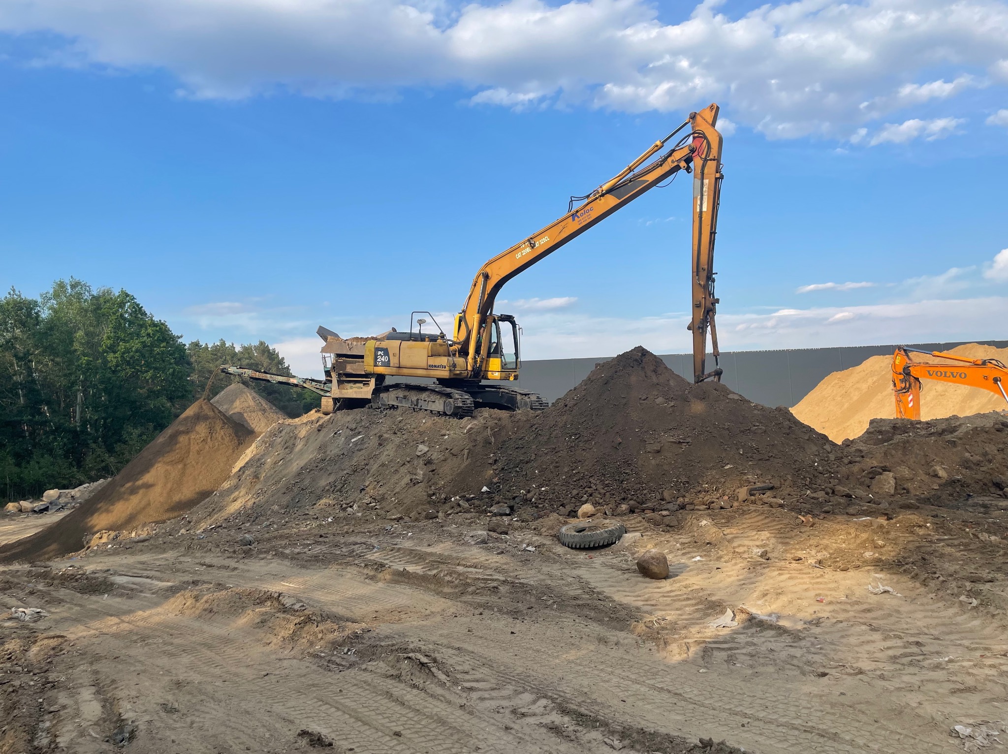 Żółta koparka Komatsu PC240 i częściowo widoczna koparka Volvo na placu budowy w Świebodzinie, z hałdami ziemi i oponą w tle, pod błękitnym niebem.