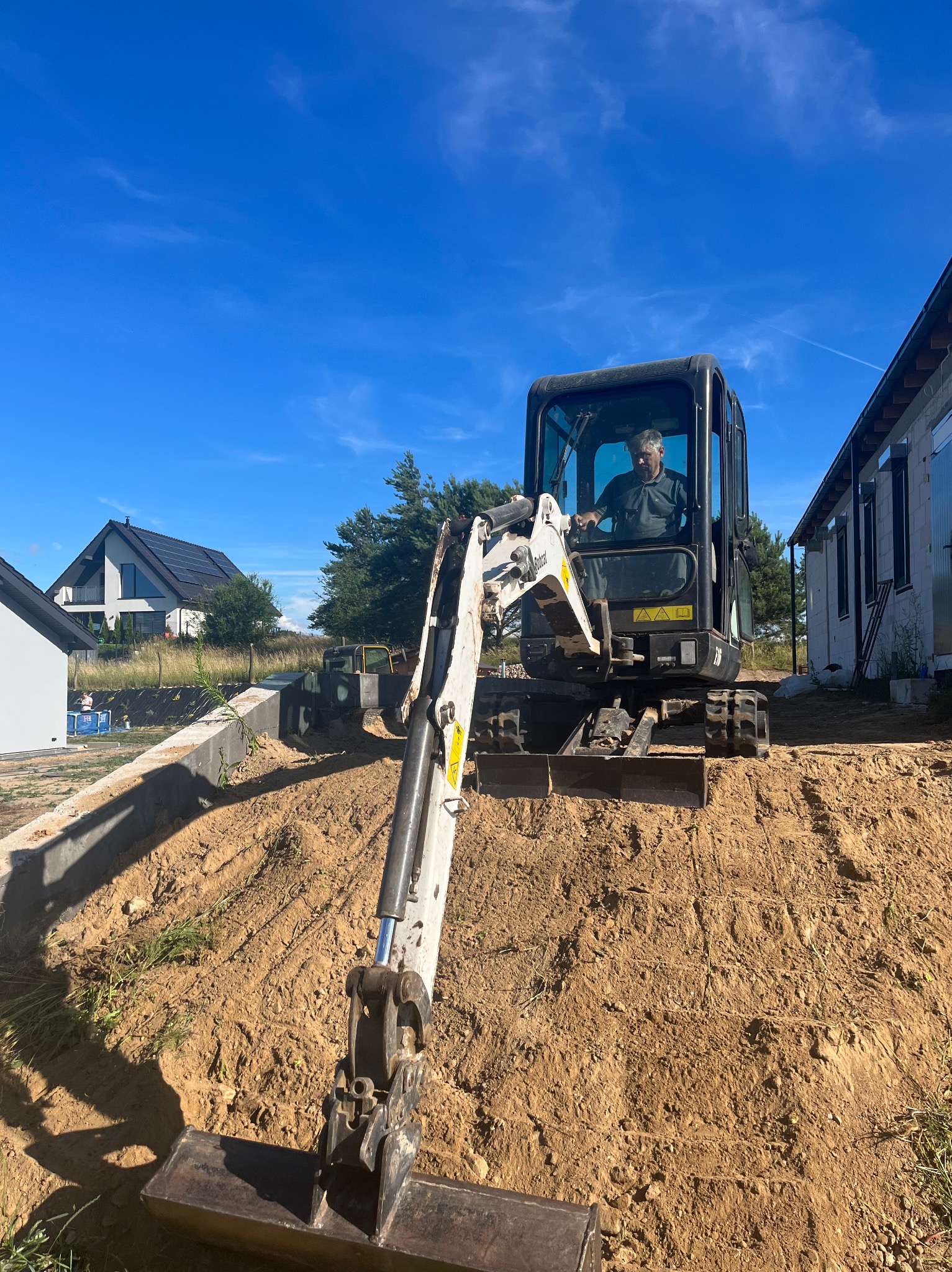 Operator w minikoparce Bobcat wyrównuje teren pod budowę domu jednorodzinnego; widoczny fragment fundamentu i sąsiednie budynki w tle, błękitne niebo.