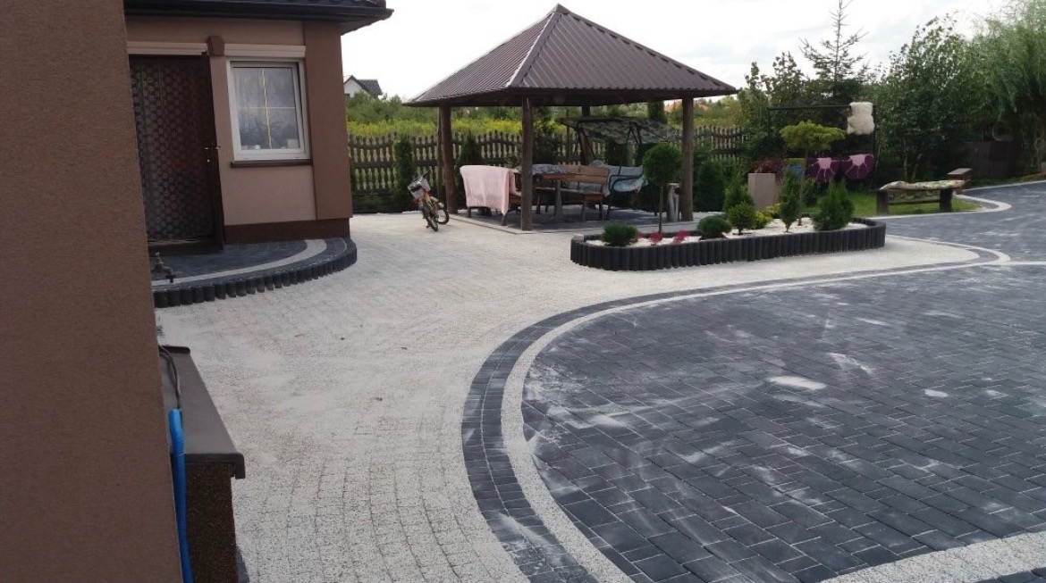 Ułożona kostka brukowa w dwóch odcieniach szarości tworząca wzór na podjeździe z altaną w ogrodzie, widoczny rower oparty o filar altany.
