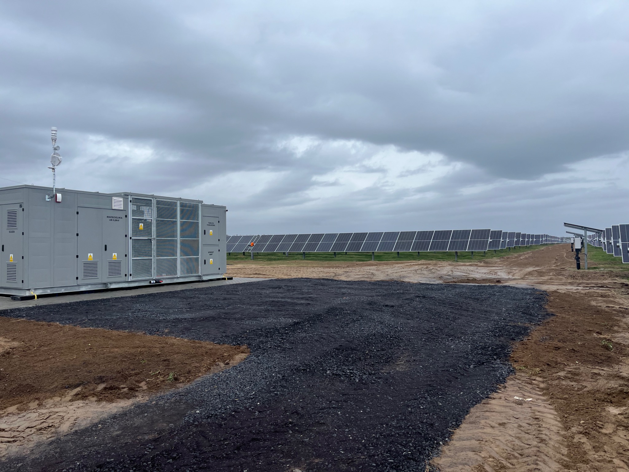 Szary kontener z instalacją elektryczną na terenie farmy fotowoltaicznej, z wysypanym świeżym, czarnym kruszywem, panele słoneczne w tle, pochmurne niebo.
