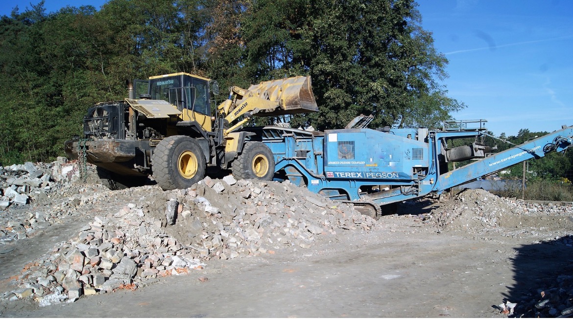 Żółta ładowarka Komatsu z łyżką podnosi gruz na niebieską kruszarkę Terex Pegson na placu budowy, w tle drzewa.
