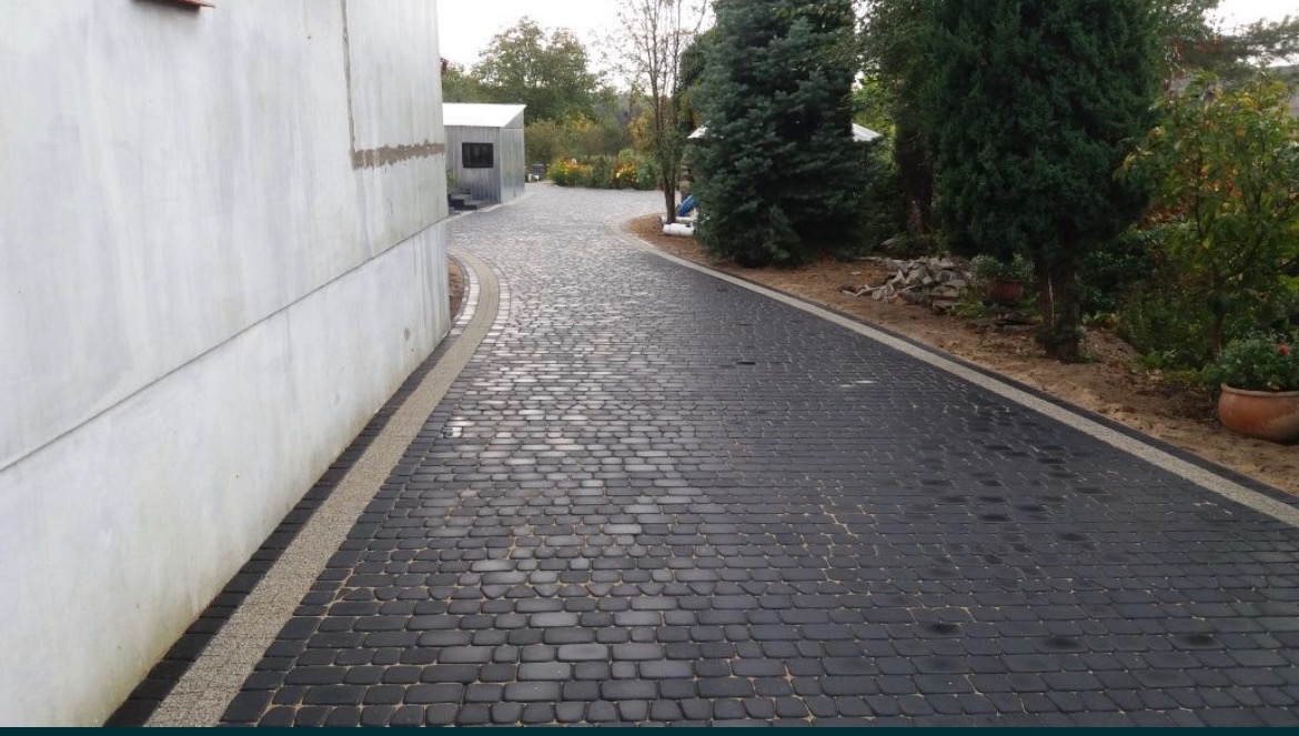 Czarna kostka brukowa ułożona na podjeździe w kształcie łuku, z jasnoszarą obwódką i budynkiem po lewej stronie oraz zielenią po prawej.