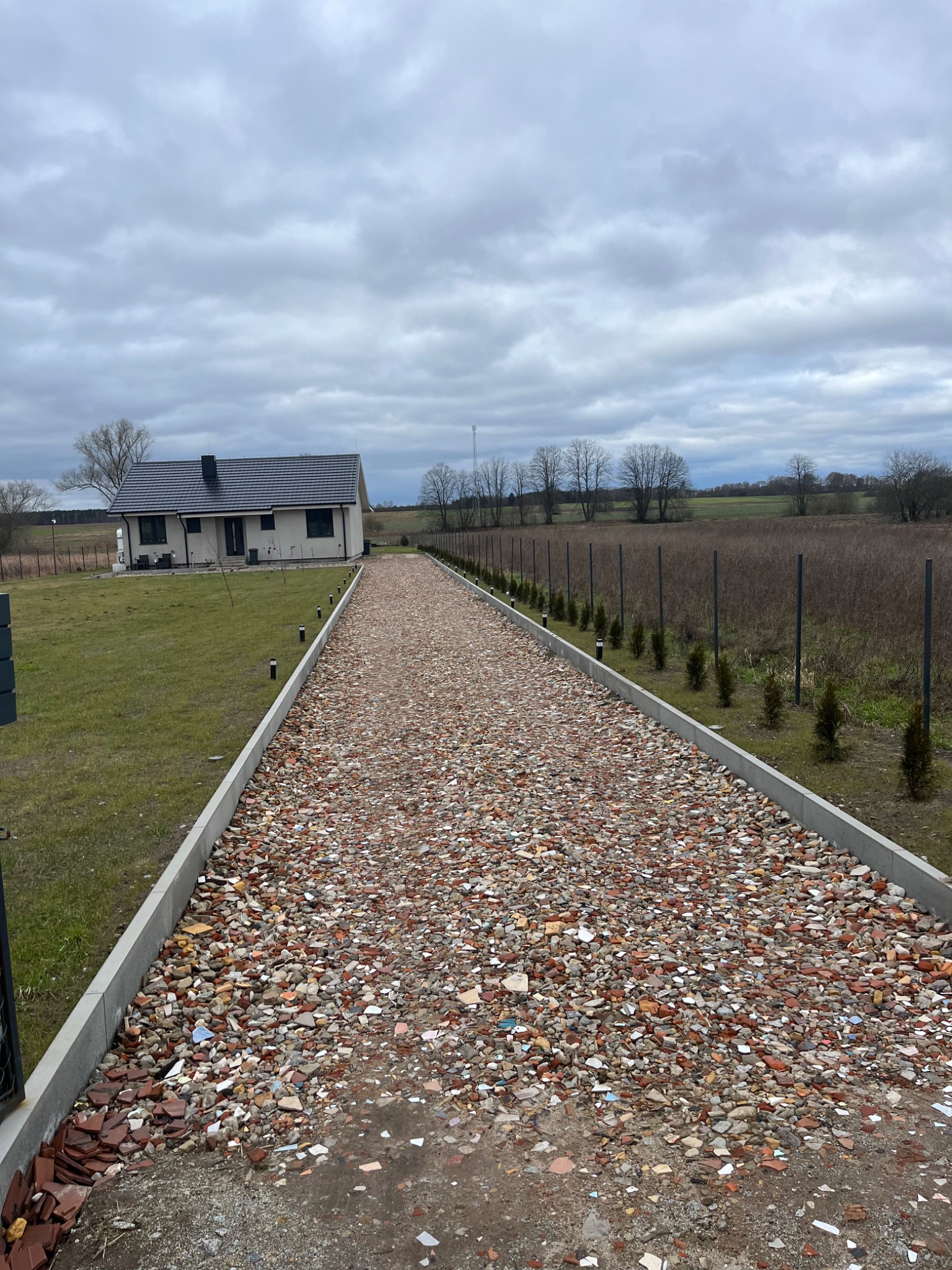 Długa, prosta droga wysypana mieszanką gruzu ceglanego i kamieni, obramowana betonowymi krawężnikami i niskimi lampami solarnymi, prowadząca do nowoczesnego domu z ciemnym dachem, widok w pochmurny...