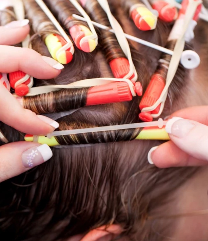 Proces trwałej ondulacji: nawijanie włosów na kolorowe wałki z gumkami zabezpieczającymi, widoczne dłonie stylistki z delikatnym manicure.