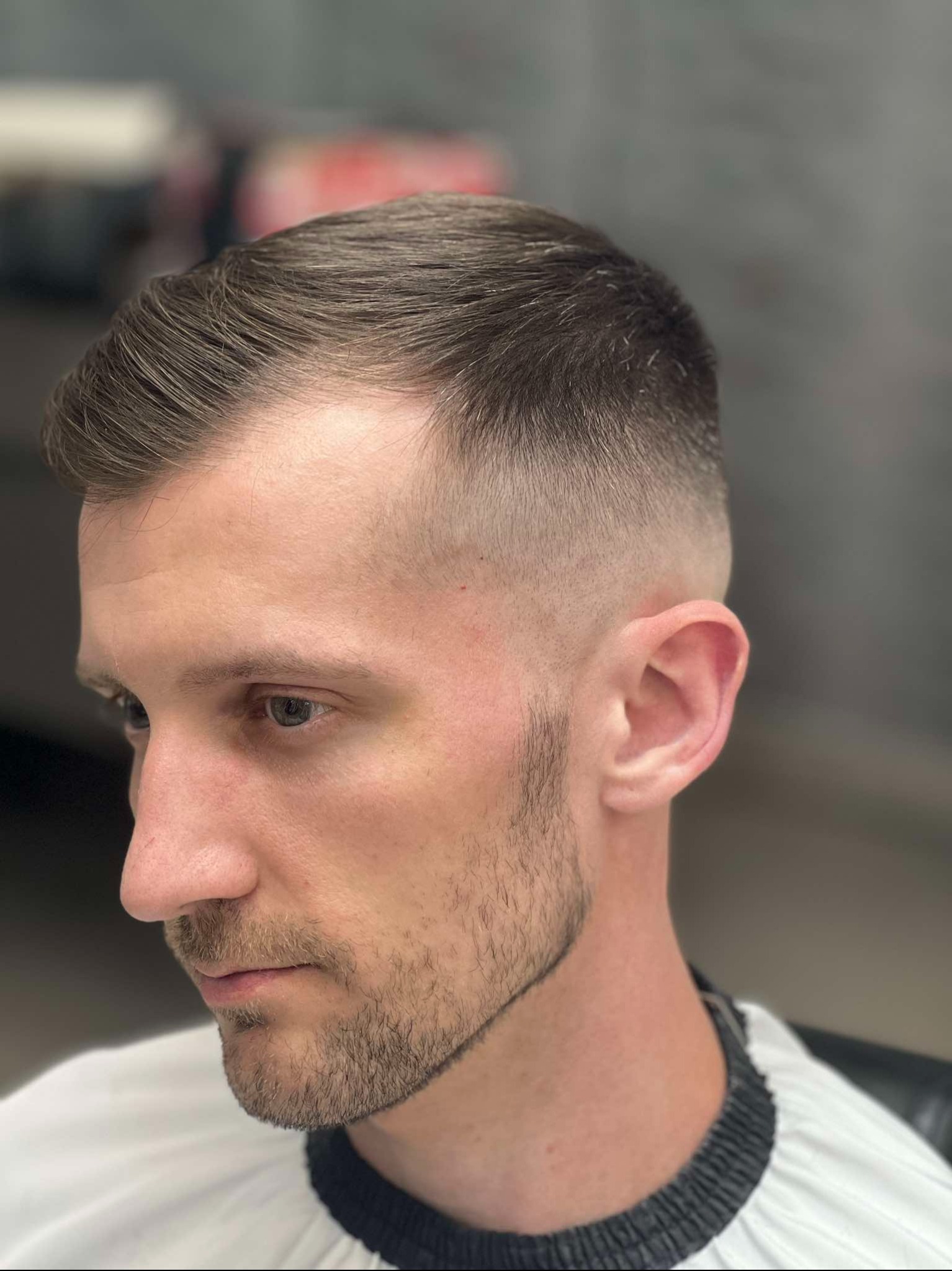 Męska fryzura z cieniowaniem, widok z profilu, zarost starannie przystrzyżony, biała peleryna fryzjerska z czarnym wykończeniem.