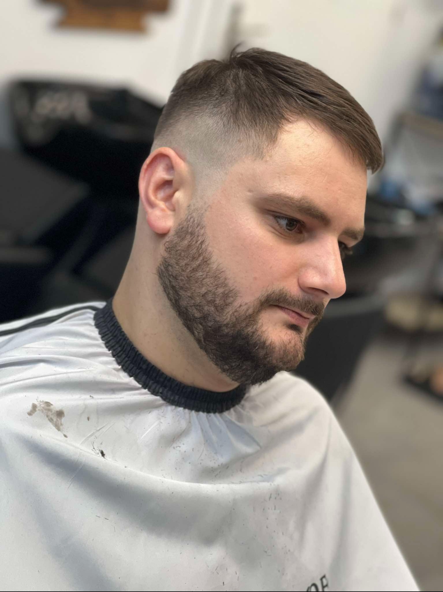 Mężczyzna z modną fryzurą fade i zadbaną brodą, profil, widoczny płaszcz ochronny z włosami po strzyżeniu.