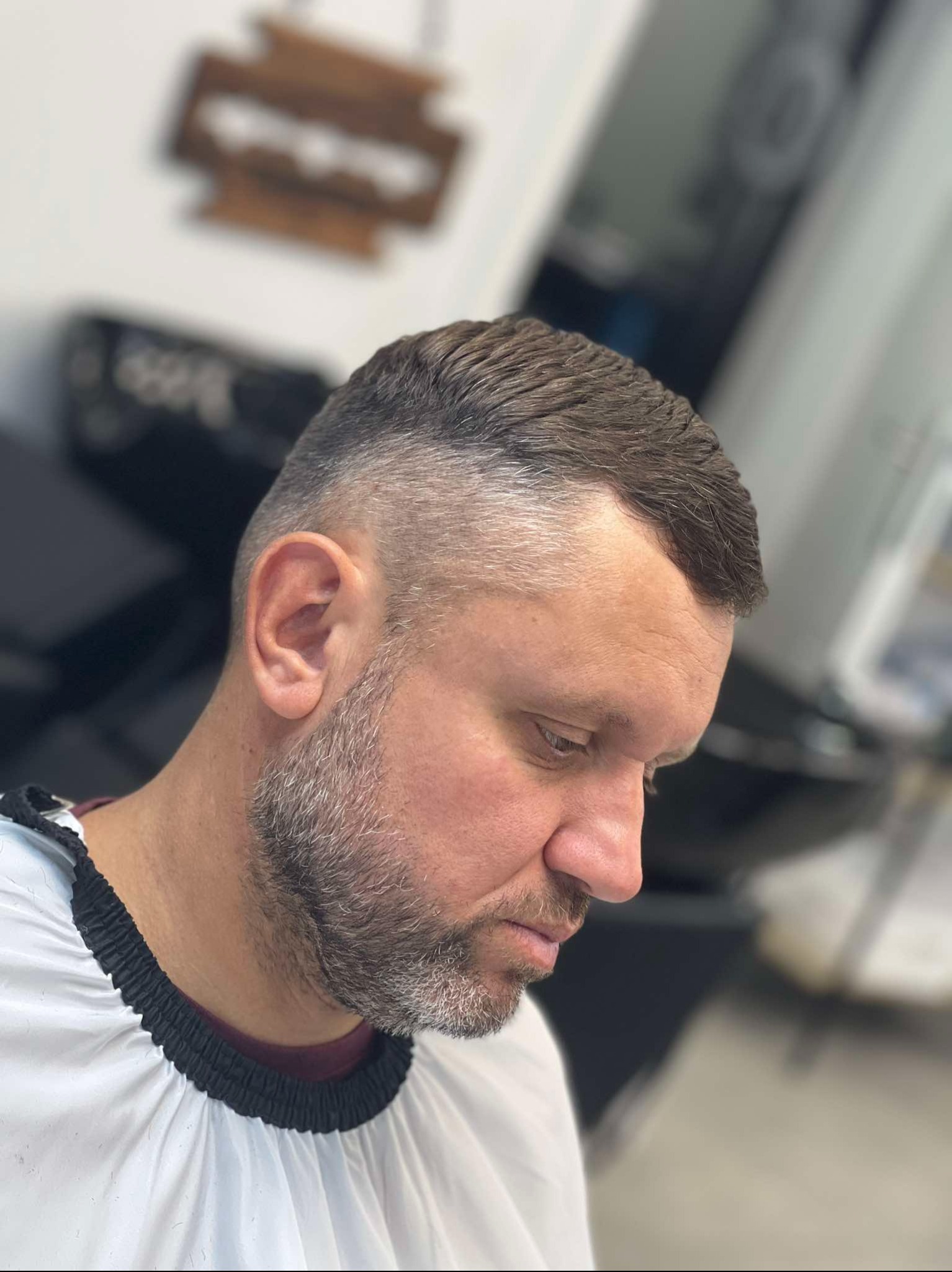 Mężczyzna z krótką fryzurą i zarostem, ubrany w białą pelerynę ochronną, spogląda w dół.