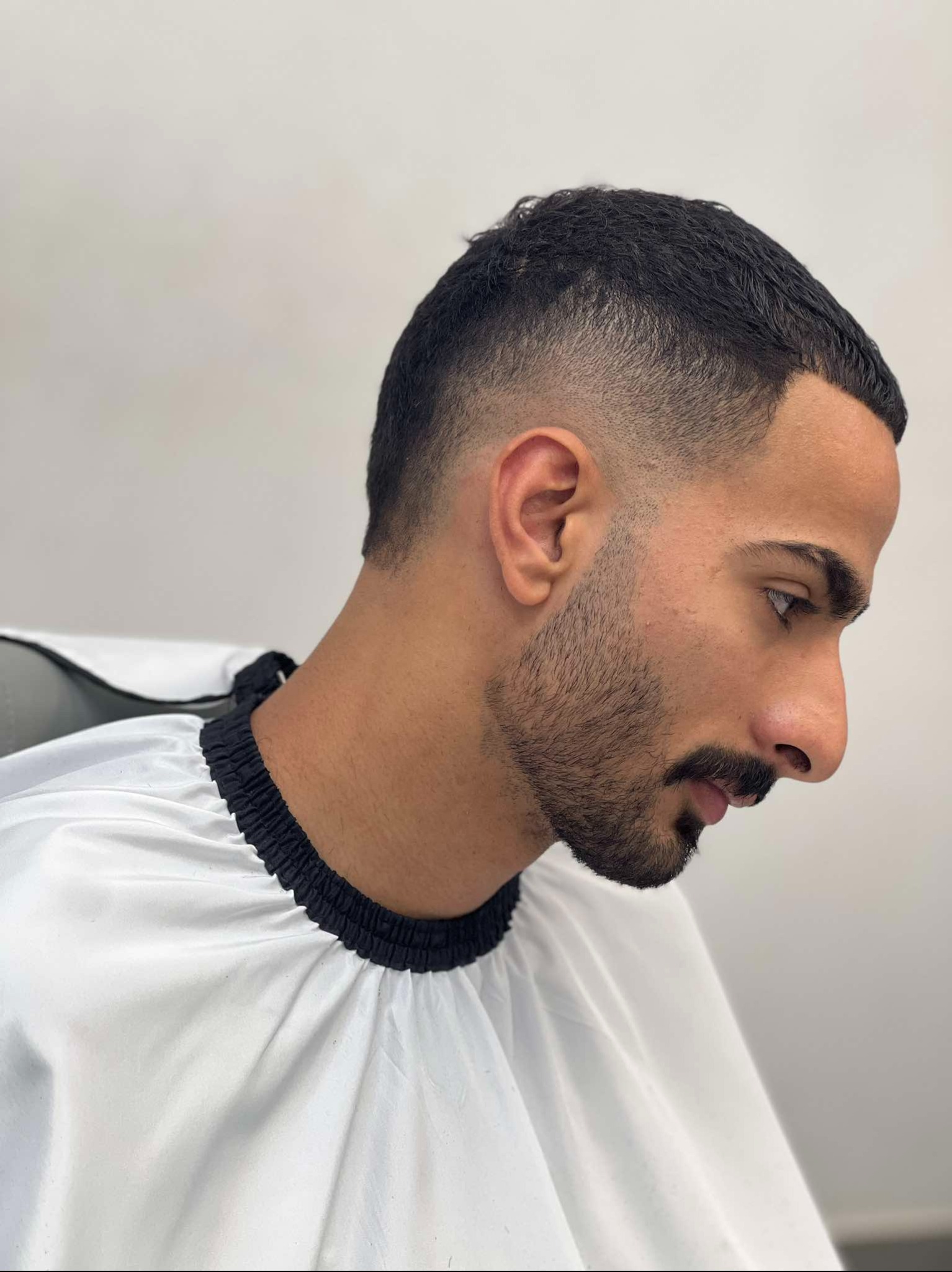 Mężczyzna z krótką, świeżo ostrzyżoną fryzurą i starannie przystrzyżonym zarostem, ubrany w białą pelerynę fryzjerską z czarnym wykończeniem, profil twarzy.