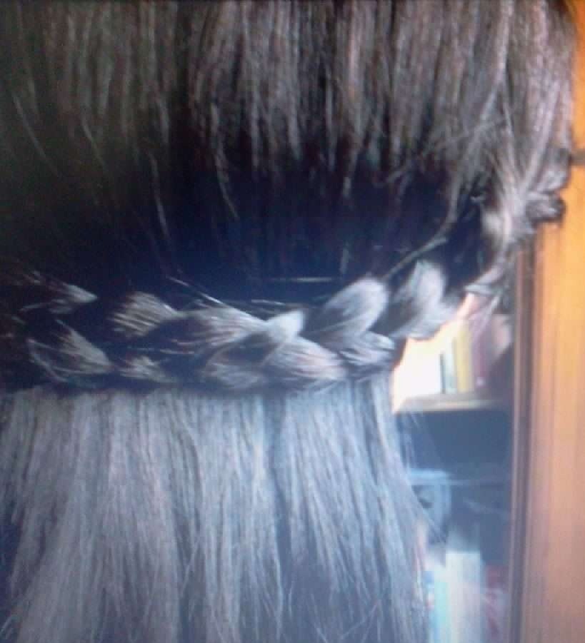 Ciemne włosy upięte w warkocz okalający głowę, widoczne od tyłu, z delikatnym zarysem półki z książkami w tle.