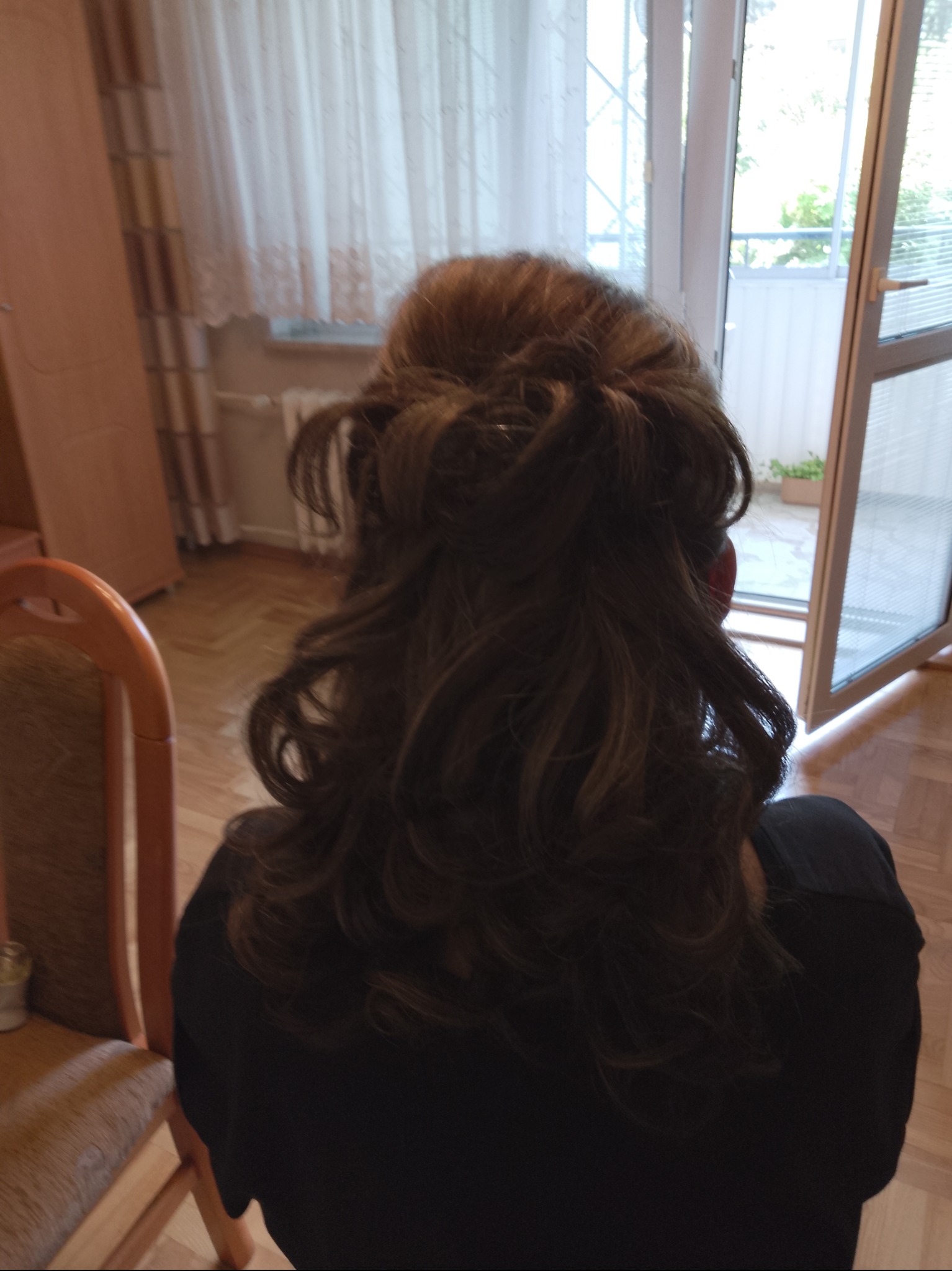 Upięcie brązowych włosów z delikatnymi falami, widok z tyłu, w tle okno balkonowe i jasne zasłony.