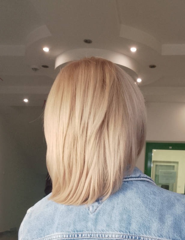 Blond włosy do ramion z tyłu, widoczne w deninowej kurtce na tle jasnego wnętrza.