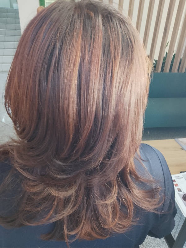 Brązowe włosy z refleksami, pocieniowane na długości, widok z tyłu, w tle jasne panele ścienne i fragment turkusowej sofy.
