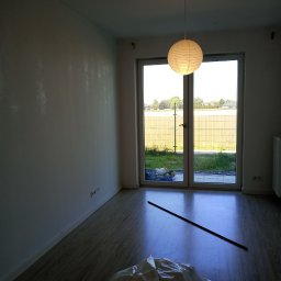 Mariusz - Pusty pokój z białymi ścianami po świeżym malowaniu, podłoga laminowana, okno balkonowe z widokiem na zewnątrz, przykryta folią podłoga i listwa przypodłogowa leżąca na podłodze.