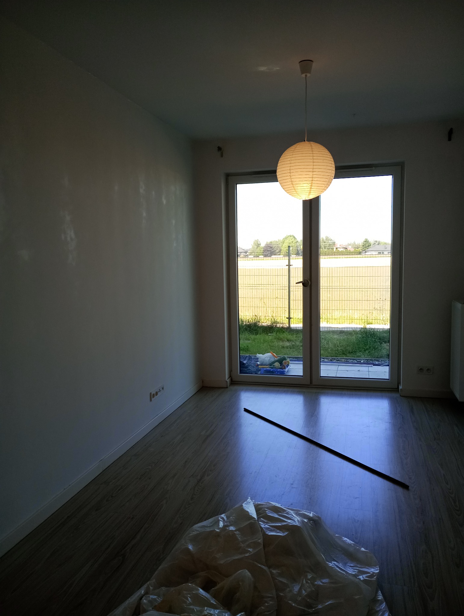 Pusty pokój z białymi ścianami po świeżym malowaniu, podłoga laminowana, okno balkonowe z widokiem na zewnątrz, przykryta folią podłoga i listwa przypodłogowa leżąca na podłodze.