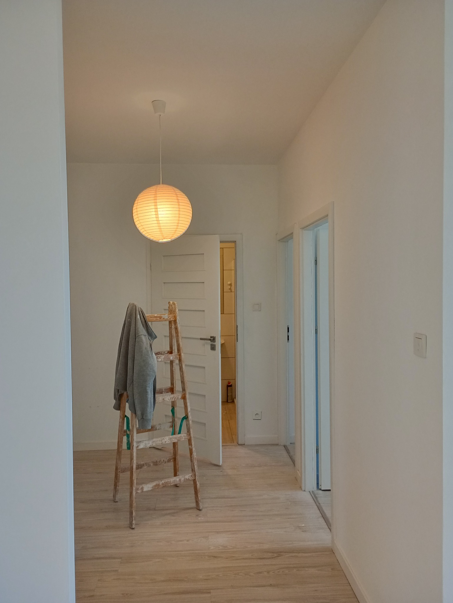 Wnętrze mieszkania po malowaniu ścian na biało, widoczna drewniana drabina z szarą bluzą i papierowa lampa wisząca.