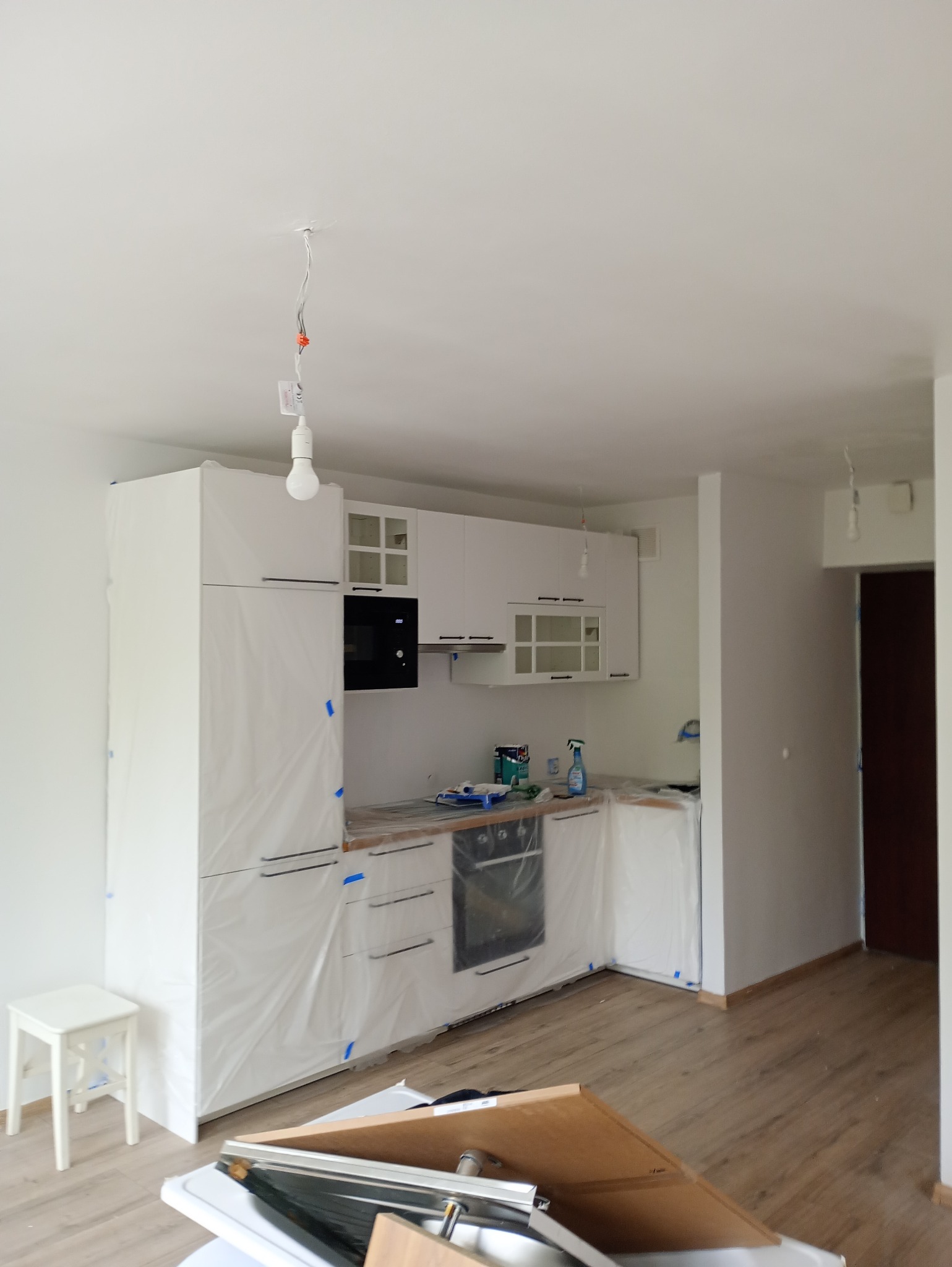 Kuchnia w trakcie przygotowań do malowania: meble oklejone folią ochronną, widoczne narzędzia malarskie, surowa żarówka zwisająca z sufitu.