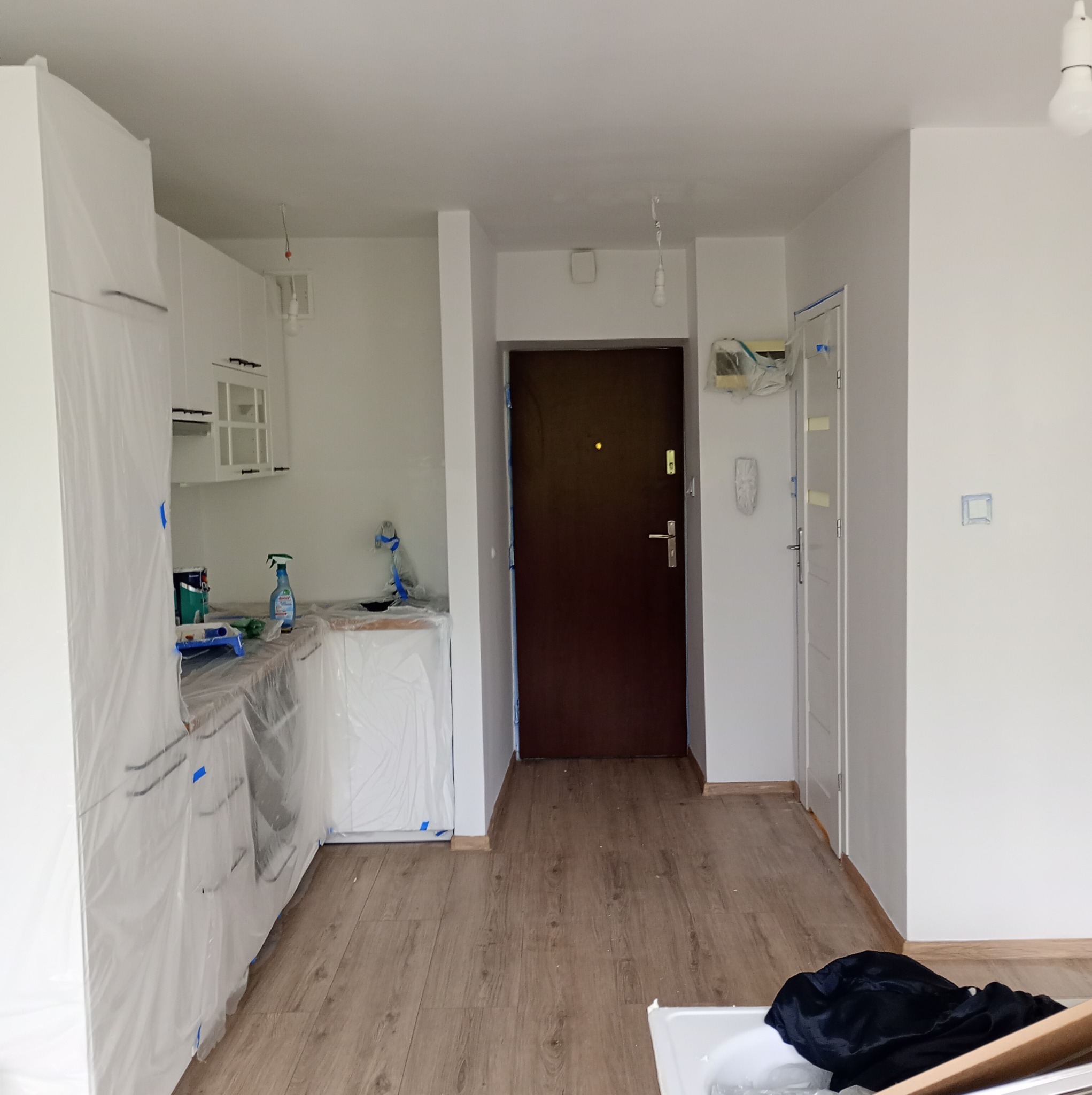 Przygotowanie do malowania mieszkania: meble kuchenne i ściany zabezpieczone folią malarską, widoczne narzędzia i środki czystości na blacie.