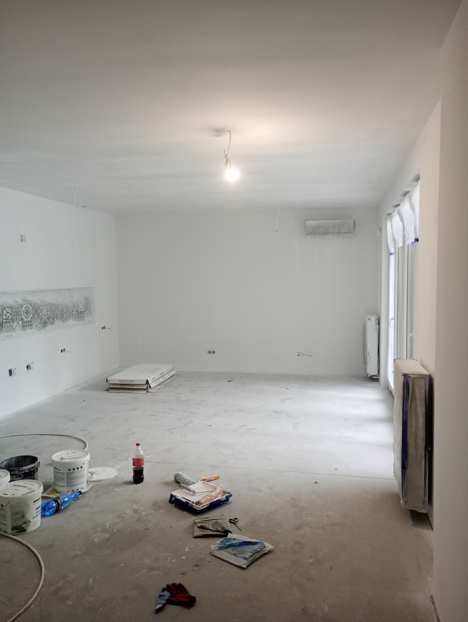 Przygotowane do malowania, białe wnętrze mieszkania z zabezpieczonymi grzejnikami i narzędziami malarskimi na podłodze.