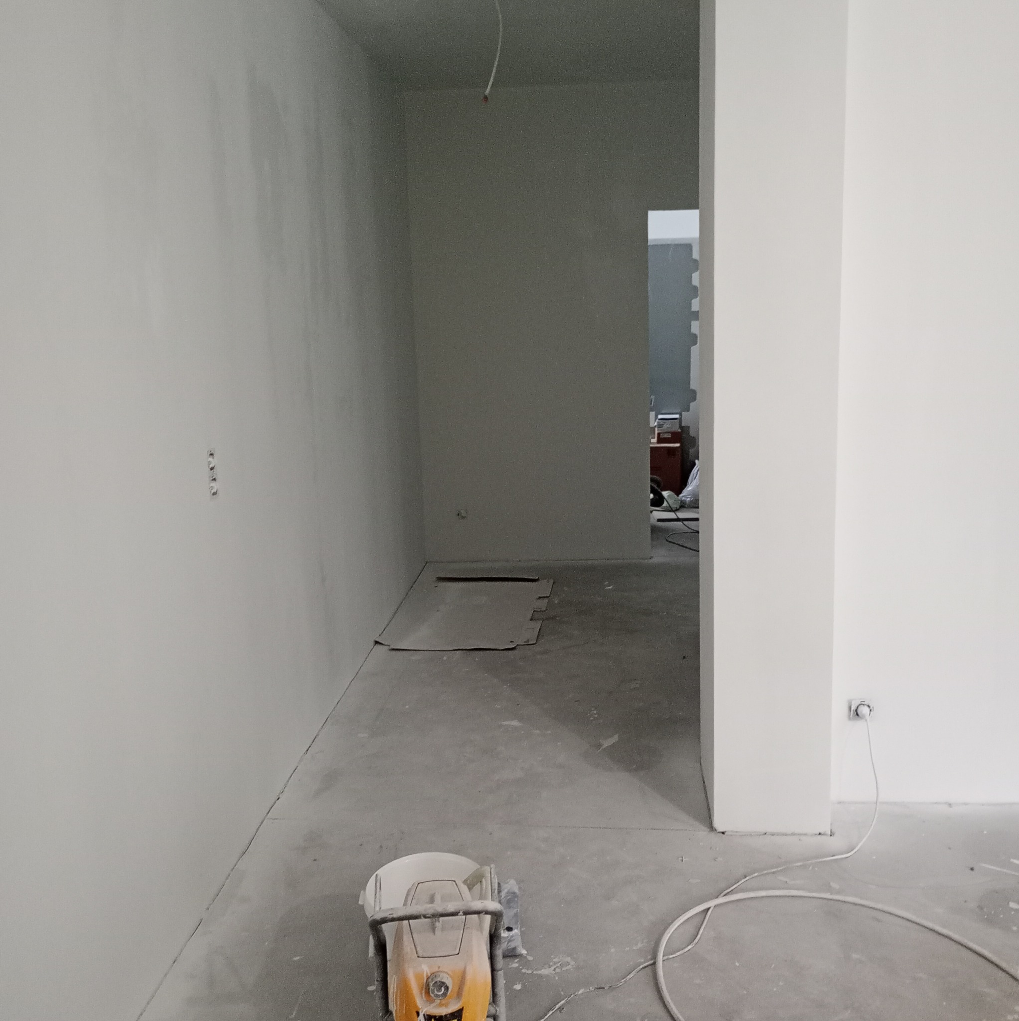 Wnętrze w trakcie remontu: świeżo wyszpachlowane ściany, szlifierka do gładzi przy podłodze, widoczne kable i gniazdko elektryczne, fragment kartonu na podłodze.