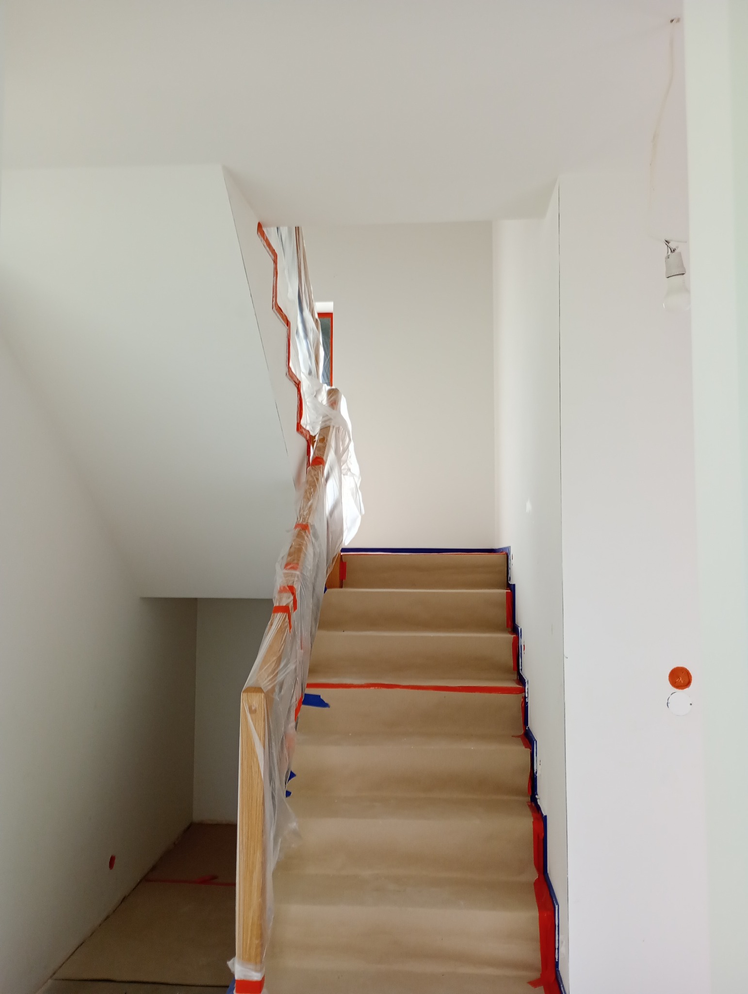 Schody zabezpieczone folią i taśmą malarską przed malowaniem ścian, widoczne pomalowane fragmenty ściany w kolorze pomarańczowym.