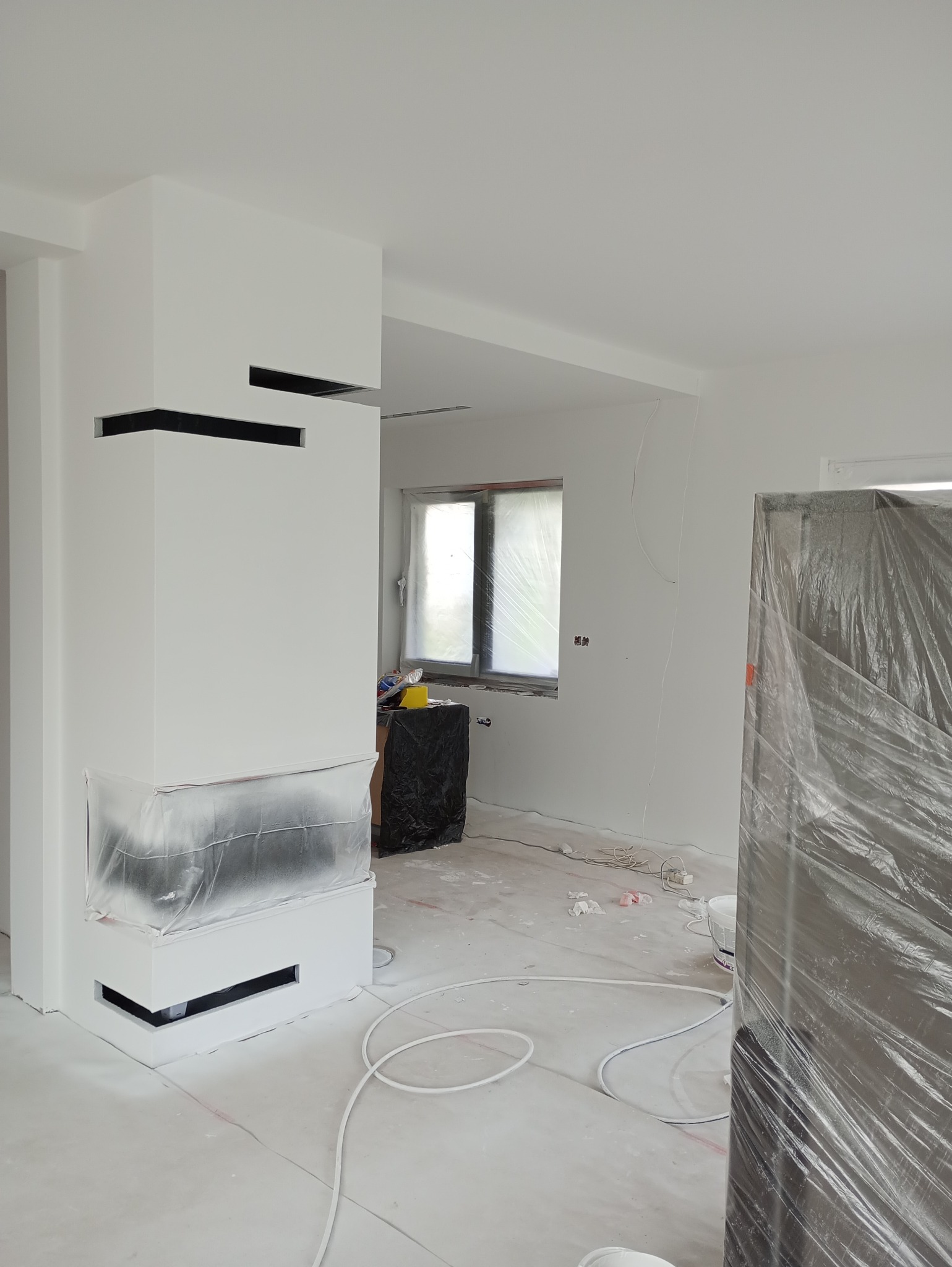 Nowoczesny kominek zabezpieczony folią malarską w trakcie prac wykończeniowych; świeżo pomalowane na biało ściany i sufit, widoczne okno z folią ochronną i kabel na podłodze.