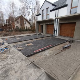 Bartosz Szymkiewicz - Prace brukarskie przed domem: ułożona szara kostka na podjeździe, czarna kostka w trakcie układania na drugim podjeździe, widoczne narzędzia i materiały budowlane, dom w tle.