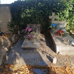 Bartosz Szymkiewicz - Dwa nagrobki z szarego kamienia otoczone kostką brukową, kwiatami i zniczami, na tle ściany porośniętej bluszczem, w słoneczny dzień.