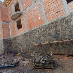 Bartosz Szymkiewicz - Wznoszenie domu z wykończoną kamienną elewacją na fundamencie, widoczne rusztowania, stosy kamieni i nieukończone mury z cegły.