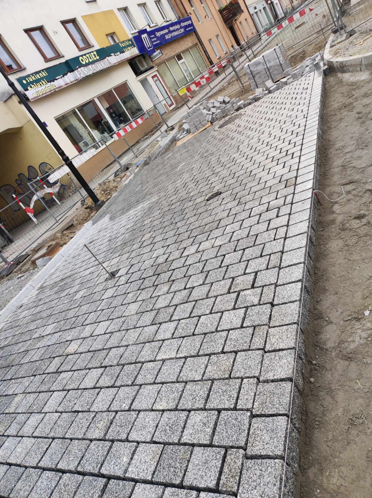 Nowa nawierzchnia z szarej kostki granitowej ułożona na ulicy miejskiej, widoczne budynki w tle i przygotowane kostki do dalszej pracy.