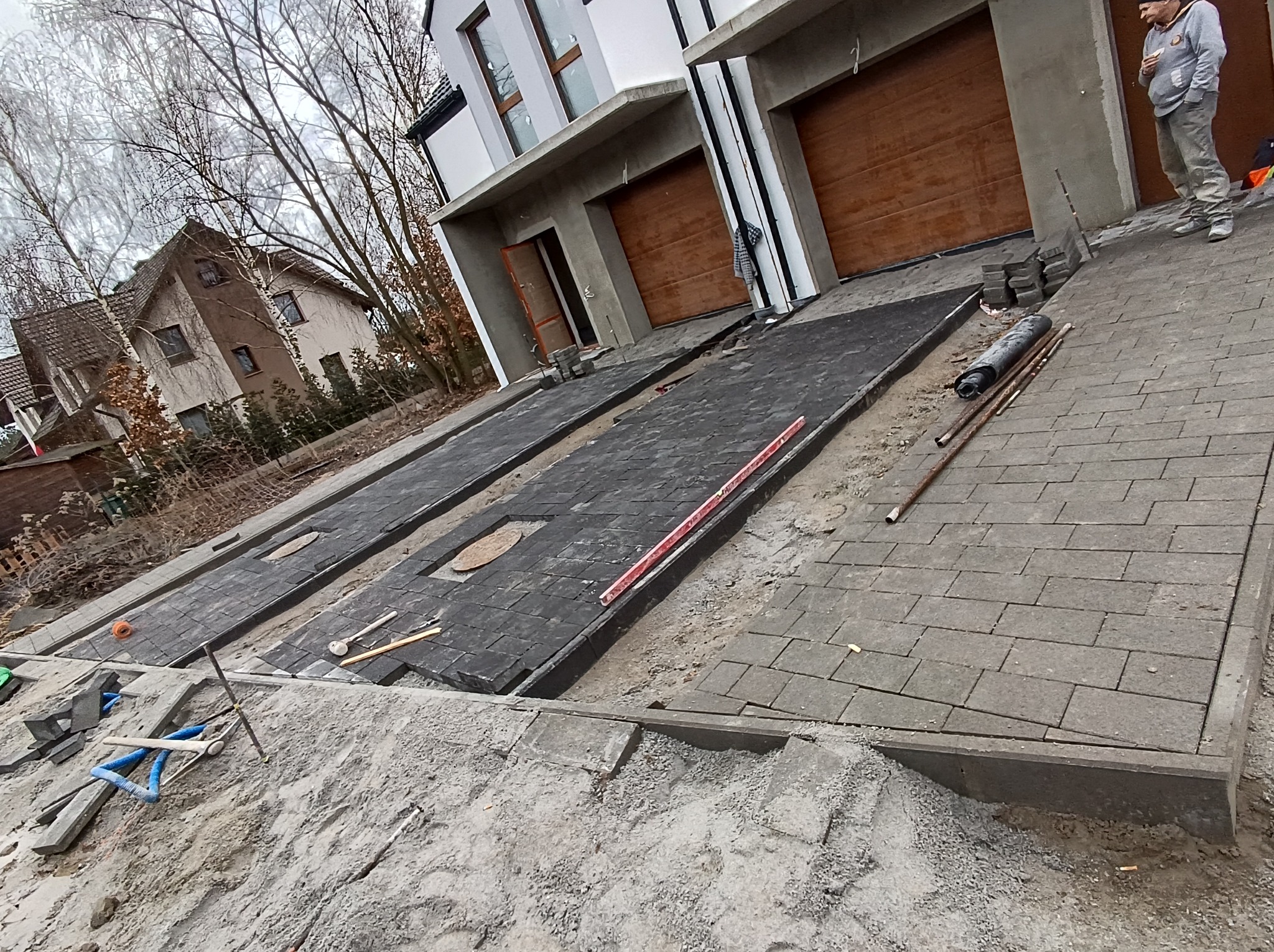 Ułożona częściowo ciemnoszara nawierzchnia przed dwoma garażami, widoczne narzędzia i materiały budowlane, w tle dom jednorodzinny i pracownik w odzieży roboczej.