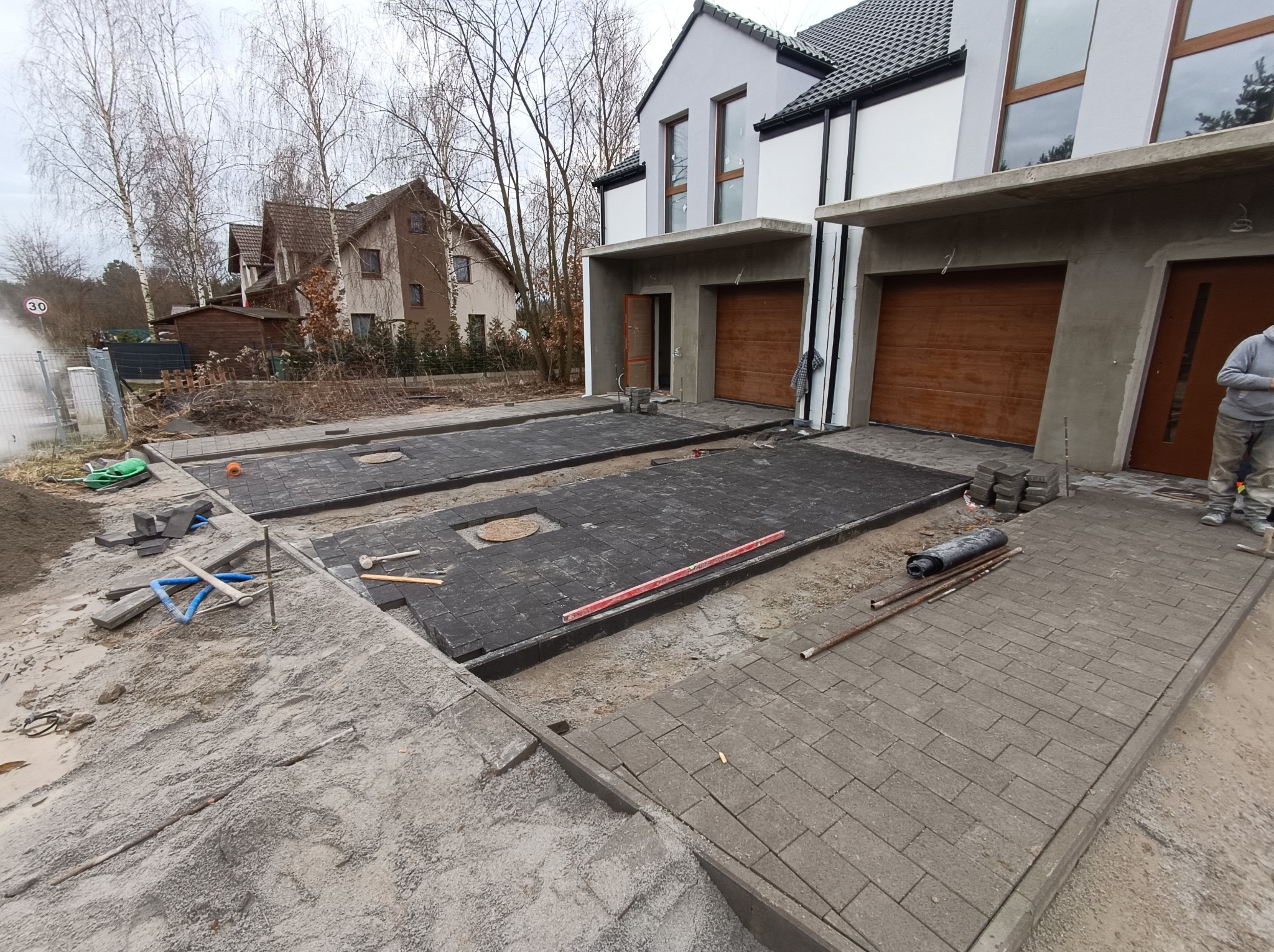 Prace brukarskie przed domem: ułożona szara kostka na podjeździe, czarna kostka w trakcie układania na drugim podjeździe, widoczne narzędzia i materiały budowlane, dom w tle.
