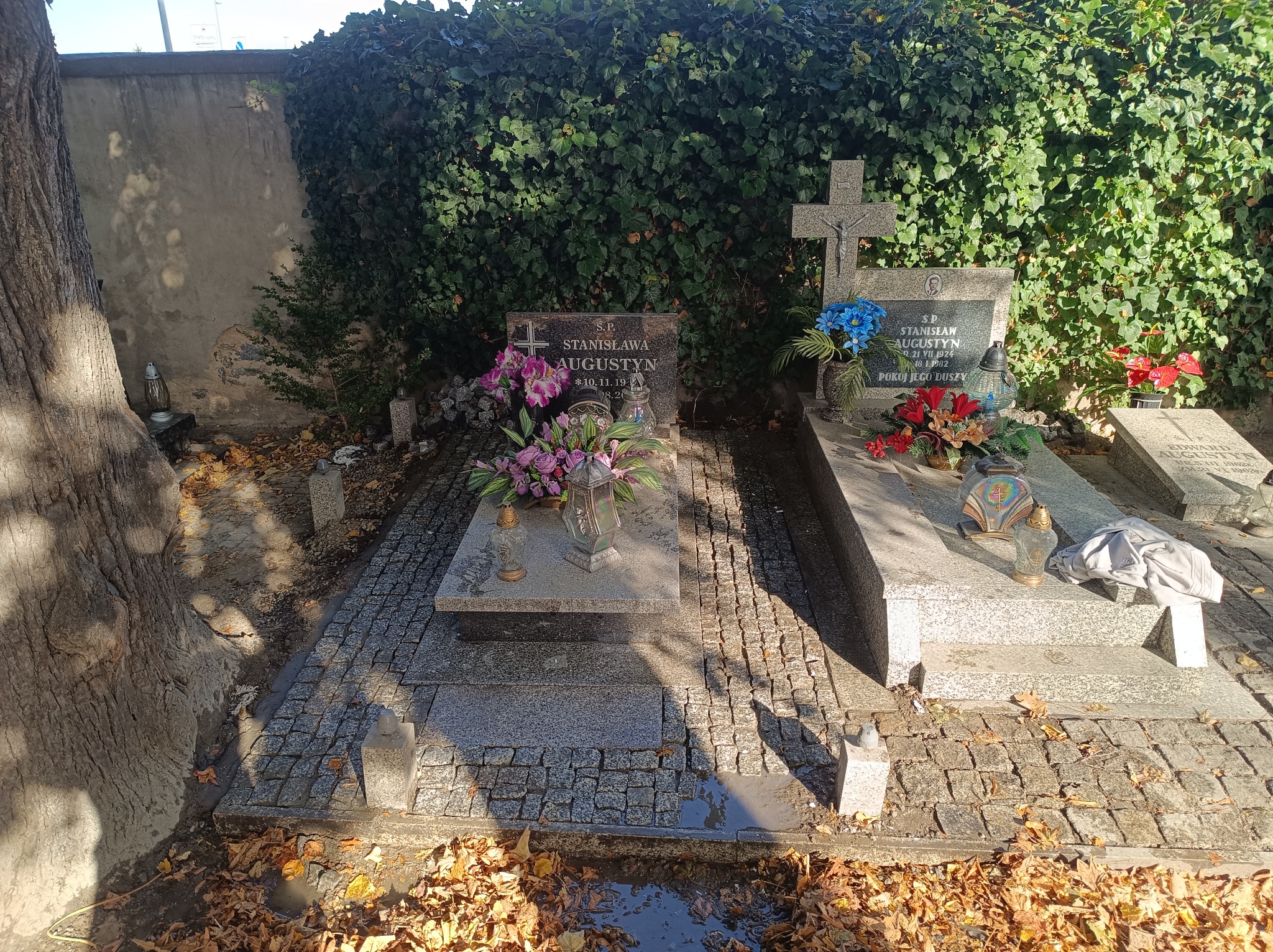 Dwa nagrobki z szarego kamienia otoczone kostką brukową, kwiatami i zniczami, na tle ściany porośniętej bluszczem, w słoneczny dzień.
