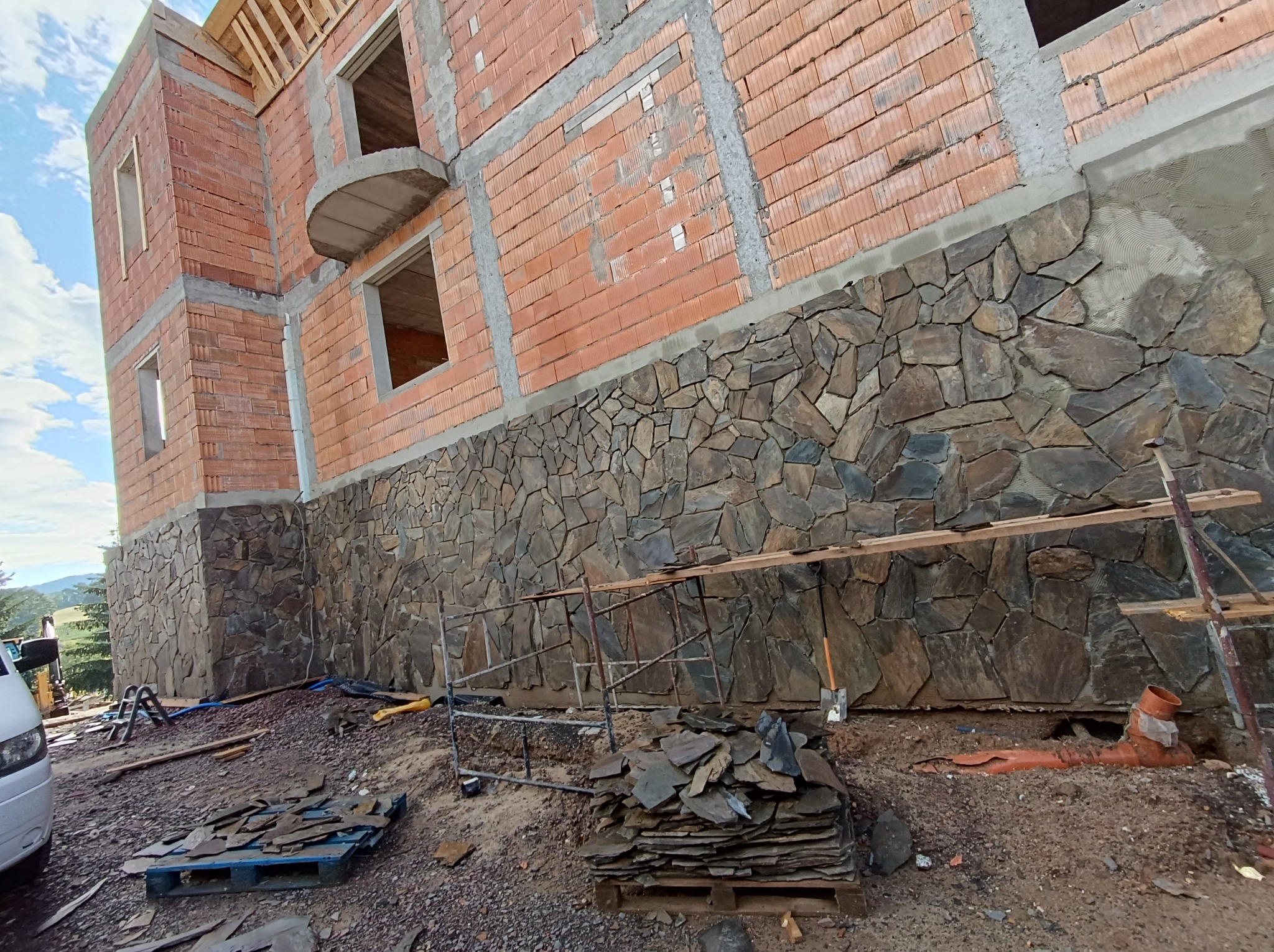 Wznoszenie domu z wykończoną kamienną elewacją na fundamencie, widoczne rusztowania, stosy kamieni i nieukończone mury z cegły.
