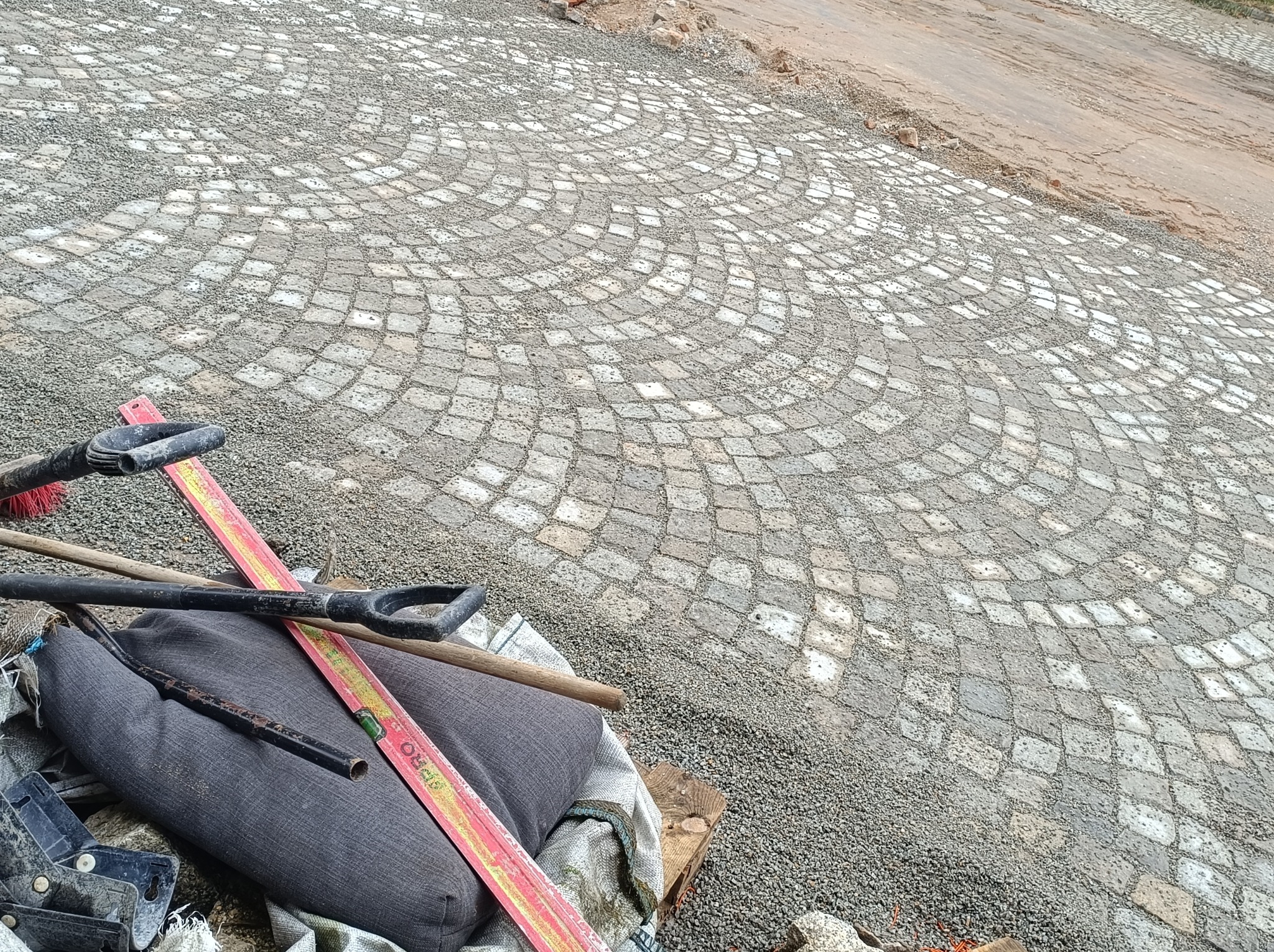 Fragment układanej nawierzchni z kostki w okrągły wzór, narzędzia brukarskie w rogu kadru, widoczny etap prac z podsypką.