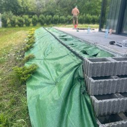 DK-TRANS - Przygotowanie rabaty z zieloną agrowłókniną, posadzone małe krzewy iglaste, w tle widoczny mężczyzna i nowoczesny dom z dużymi oknami.