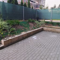 DK-TRANS - Ułożona kostka brukowa na podwórku z niskim murkiem, za którym widać roślinność i zieloną siatkę ogrodzeniową. Częściowo widoczne rusztowania w tle.