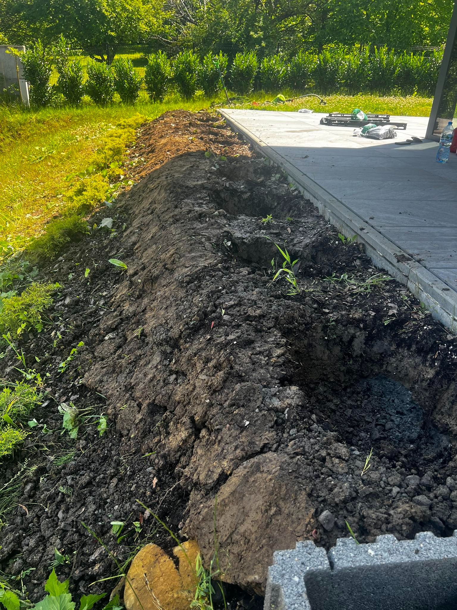 Przygotowany pas ziemi obok tarasu, gotowy do sadzenia roślin, z widocznymi narzędziami ogrodniczymi w tle.