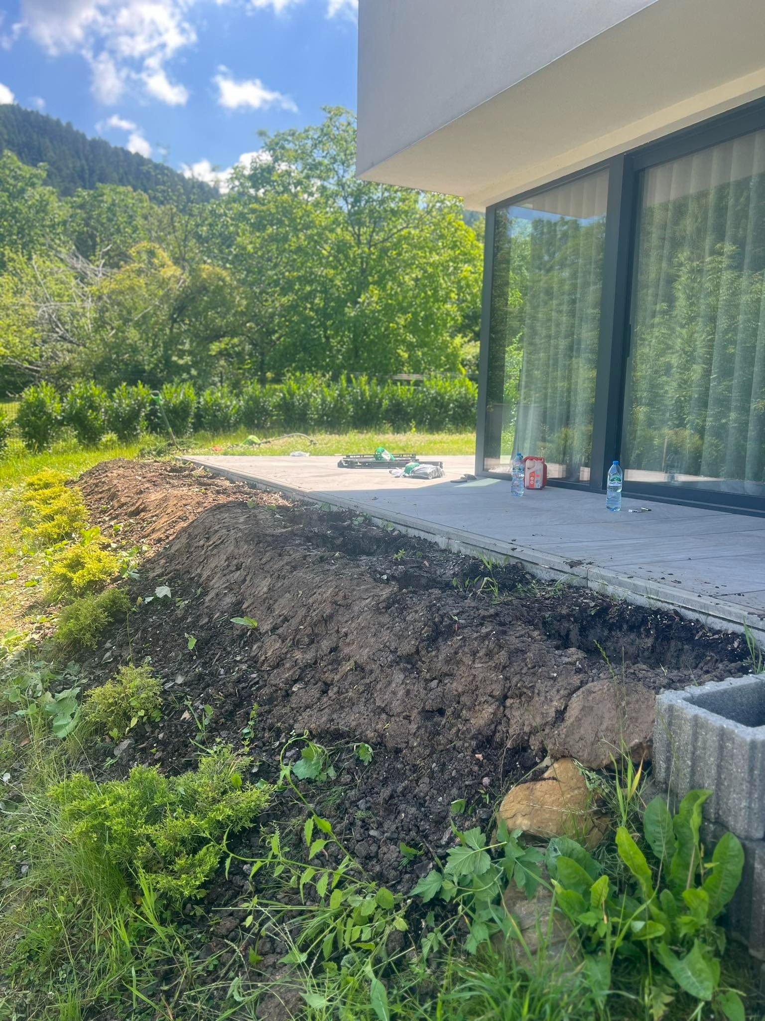Przygotowany pas ziemi pod nasadzenia wzdłuż tarasu nowoczesnego domu, widoczne narzędzia ogrodnicze i butelki wody na tarasie, w tle zalesione wzgórza.