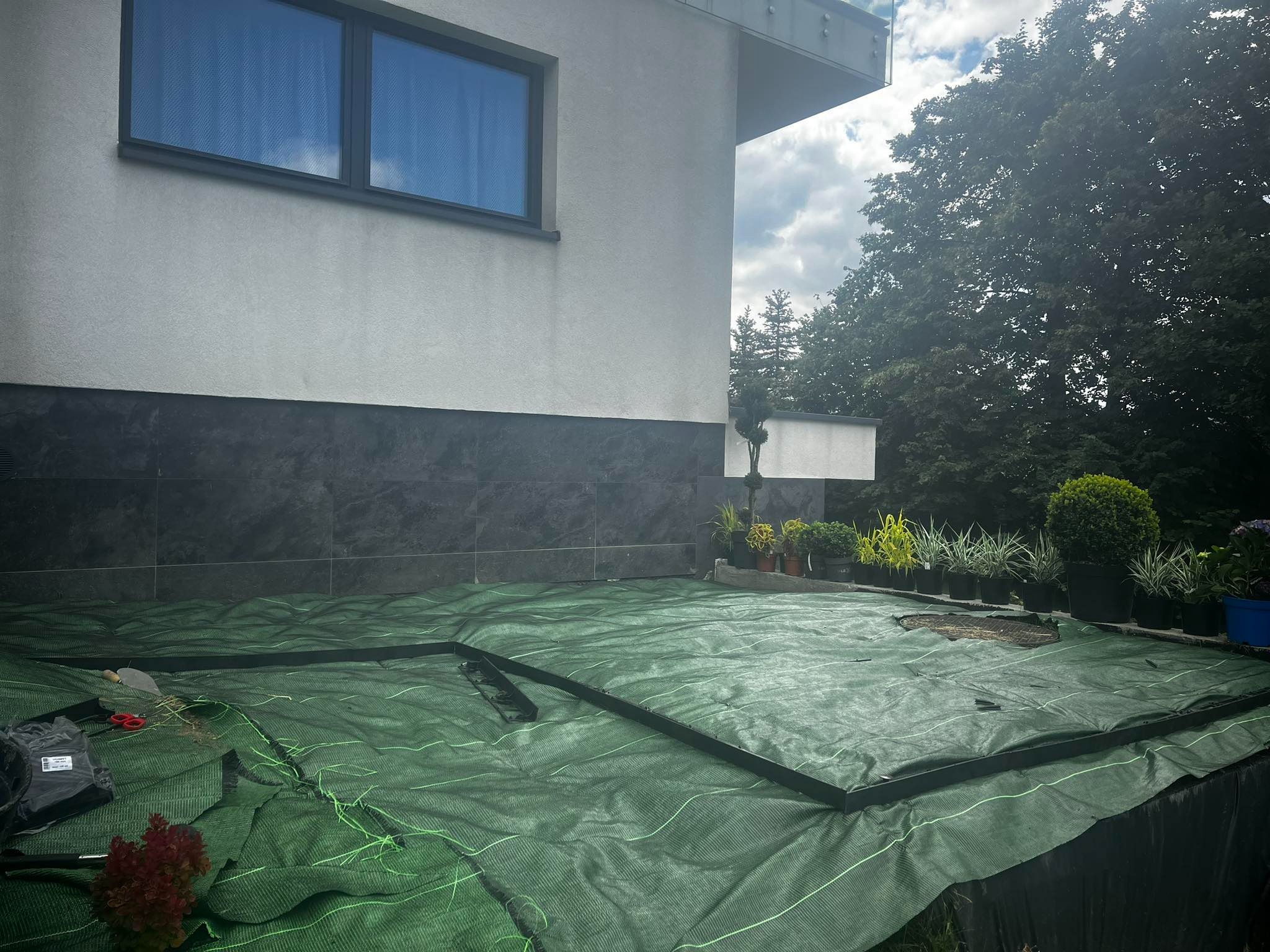 Przygotowanie terenu pod nasadzenia w ogrodzie: rozłożona zielona agrowłóknina z widocznymi narzędziami ogrodniczymi, obrzeżami trawnikowymi i młodymi roślinami w doniczkach w tle, na tle ściany...