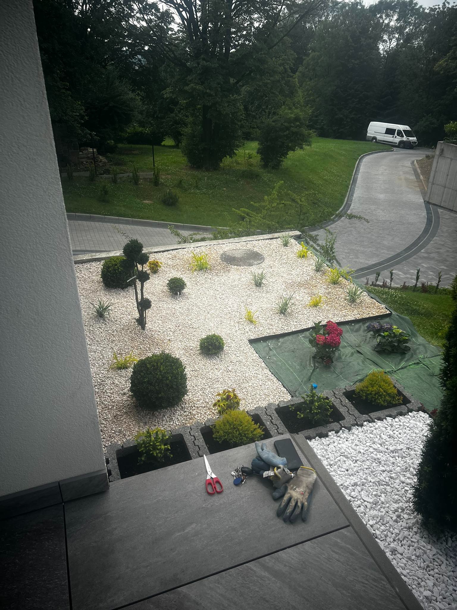 Nowo założony ogród z białym żwirem, roślinami w donicach z betonu, narzędziami ogrodniczymi i samochodem dostawczym w tle.