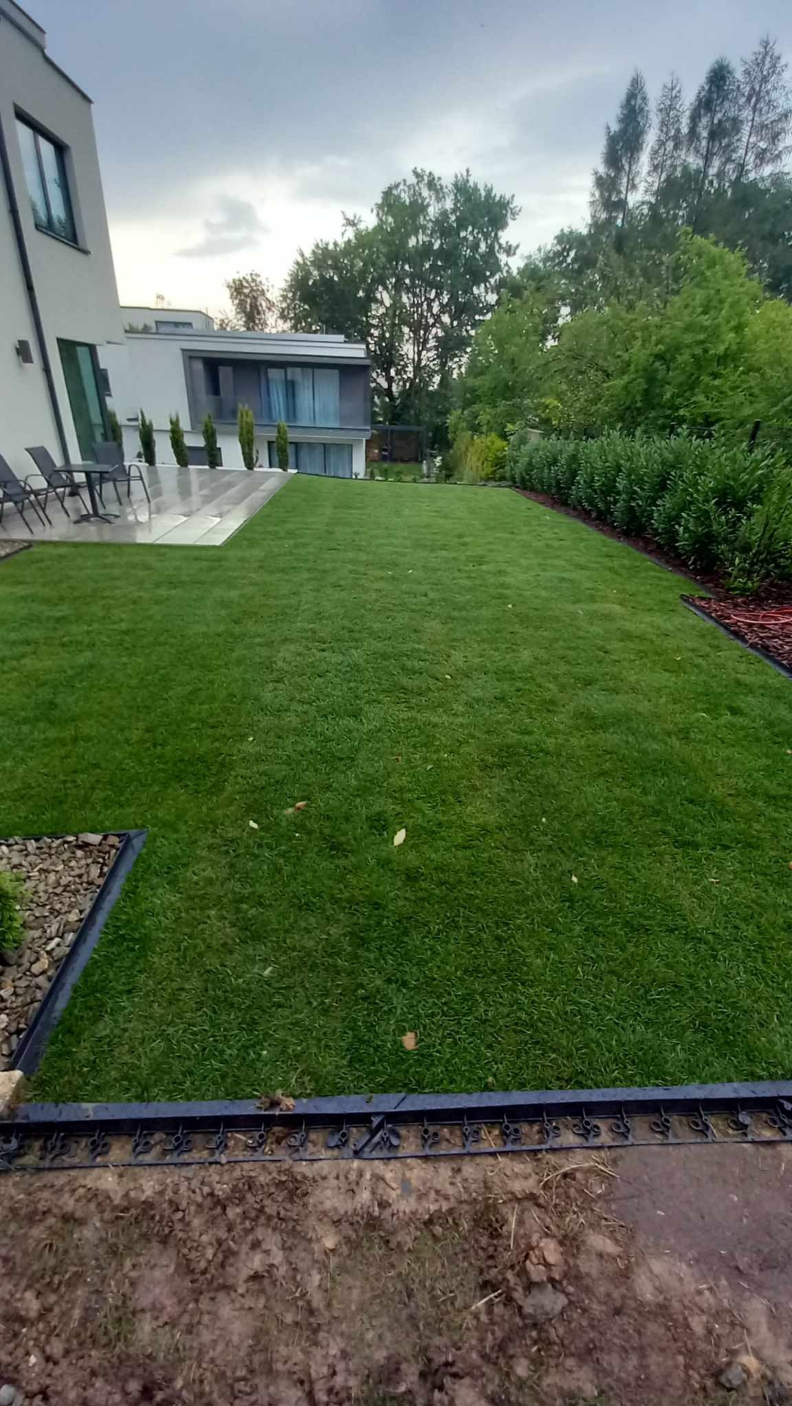 Idealnie przystrzyżony trawnik przed nowoczesnym domem z tarasem i meblami ogrodowymi, obrzeża trawnika wykończone estetyczną bordiurą i korą.