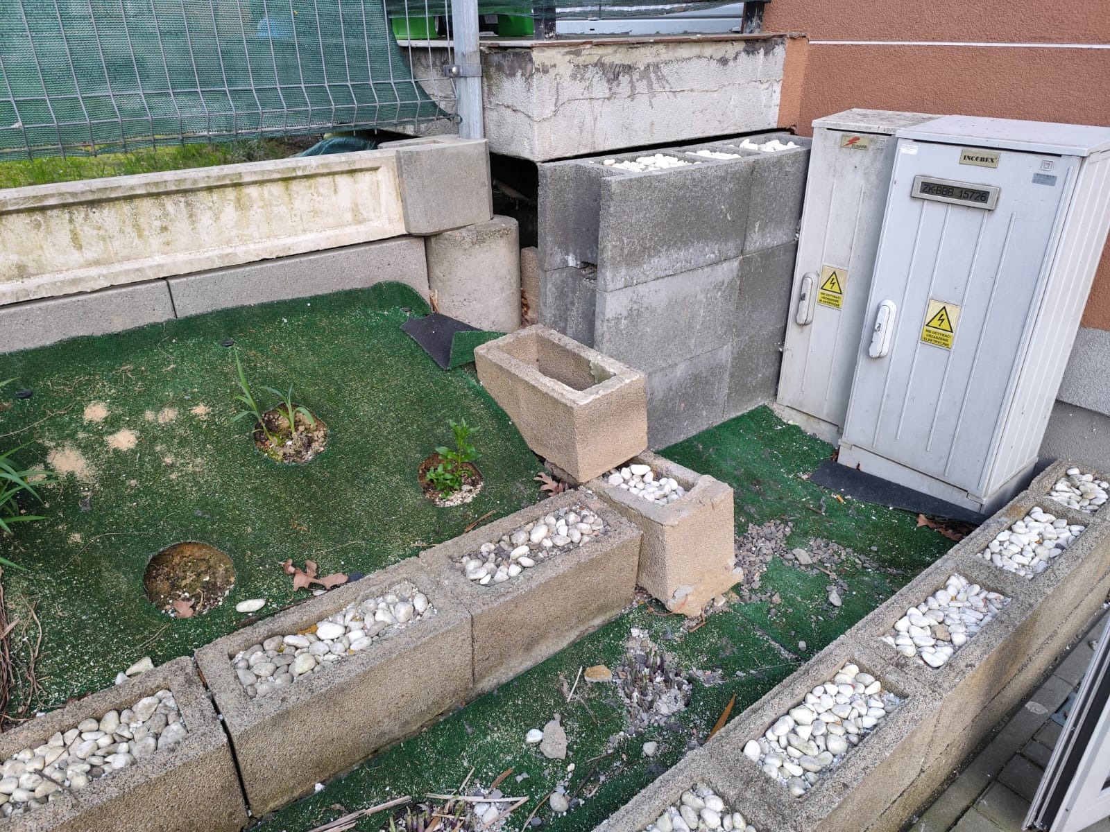 Niedokończony ogródek z betonowych bloczków wypełnionych białymi kamieniami, sztuczna trawa, skrzynka elektryczna przy ścianie budynku.