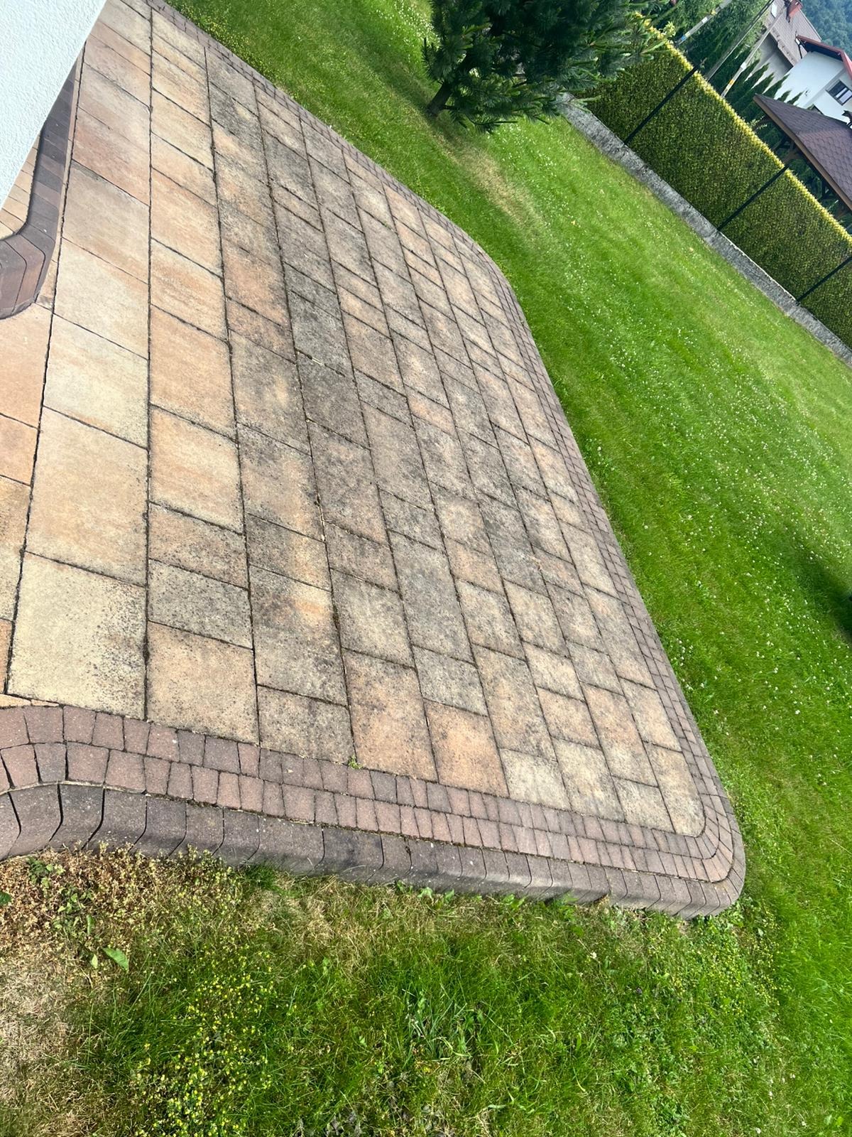 Wyraźny widok na kostkę brukową po czyszczeniu, gdzie widoczna jest różnica w kolorze i czystości w porównaniu do stanu pierwotnego. Trawa wokół kostki jest zielona i zadbana.