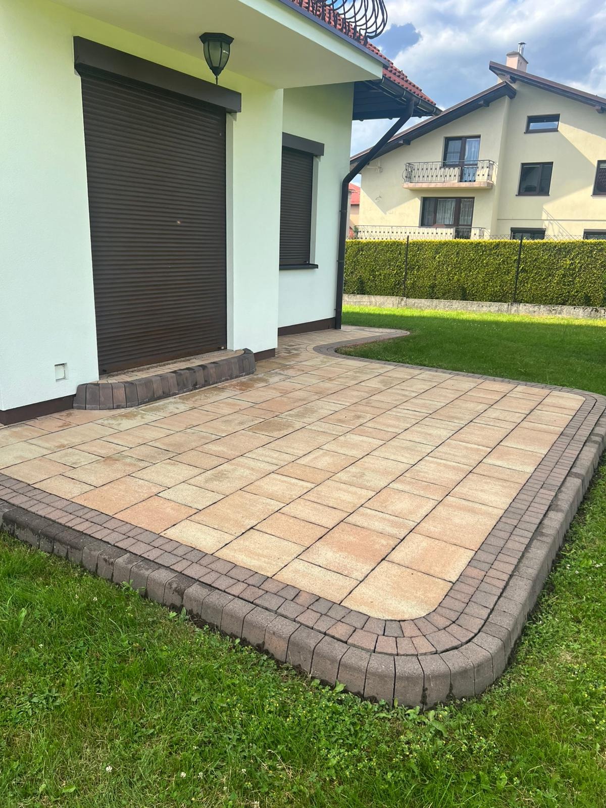 Ułożona kostka brukowa w odcieniach beżu i brązu tworzy estetyczny taras przy domu z zamkniętymi, brązowymi roletami. Zadbany trawnik otacza nawierzchnię.