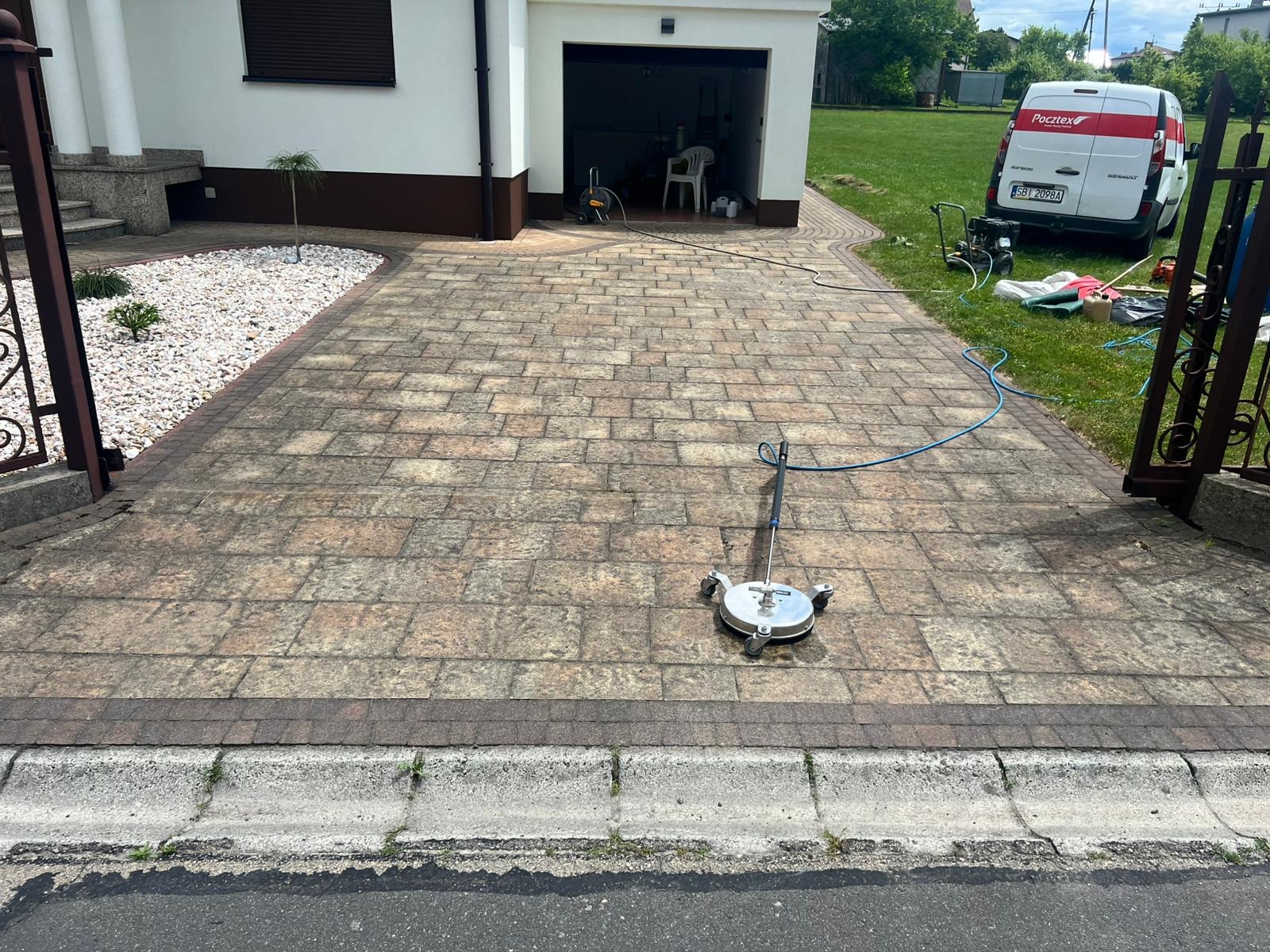Mycie kostki brukowej przy użyciu rotacyjnej myjki ciśnieniowej. Widoczny fragment posesji z kostką, brama, trawnik i budynek w tle. Wąż doprowadzający wodę.