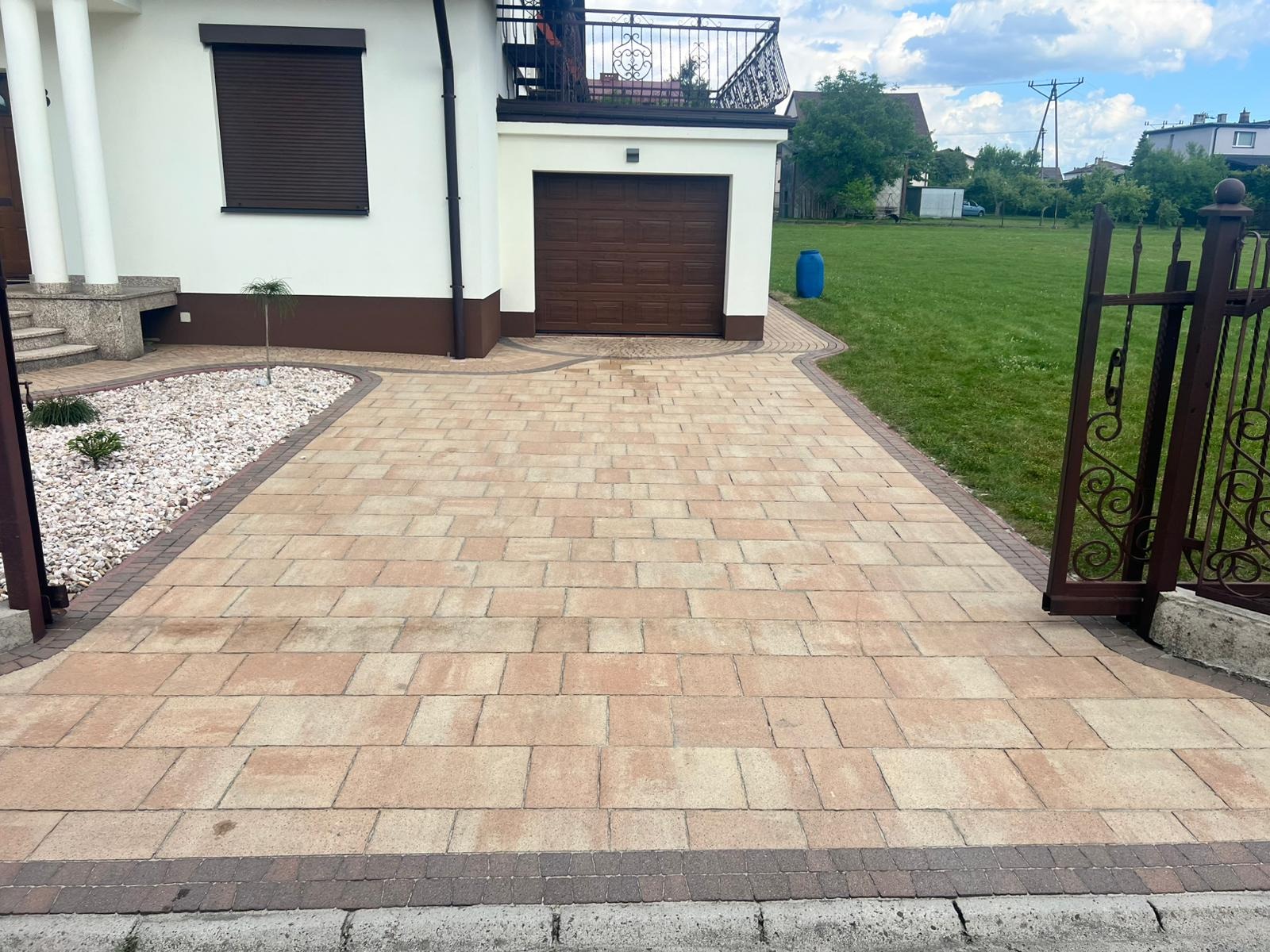 Ułożona kostka brukowa w odcieniach beżu i brązu tworzy wzorzysty podjazd do domu z garażem. Widoczne elementy ogrodu i brama wjazdowa.