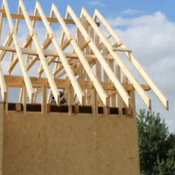 Wznoszenie drewnianej konstrukcji domu: ściany z płyt OSB i szkielet dachu na tle błękitnego nieba z chmurami.