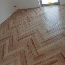 PROFESJONALNE WYKOŃCZENIE WNĘTRZ RAFMAG RAFAŁ PIERNICKI - Ułożona podłoga z paneli w jodełkę, widok z góry, jasne drewno, okno w tle. Nowoczesne wykończenie poddasza.