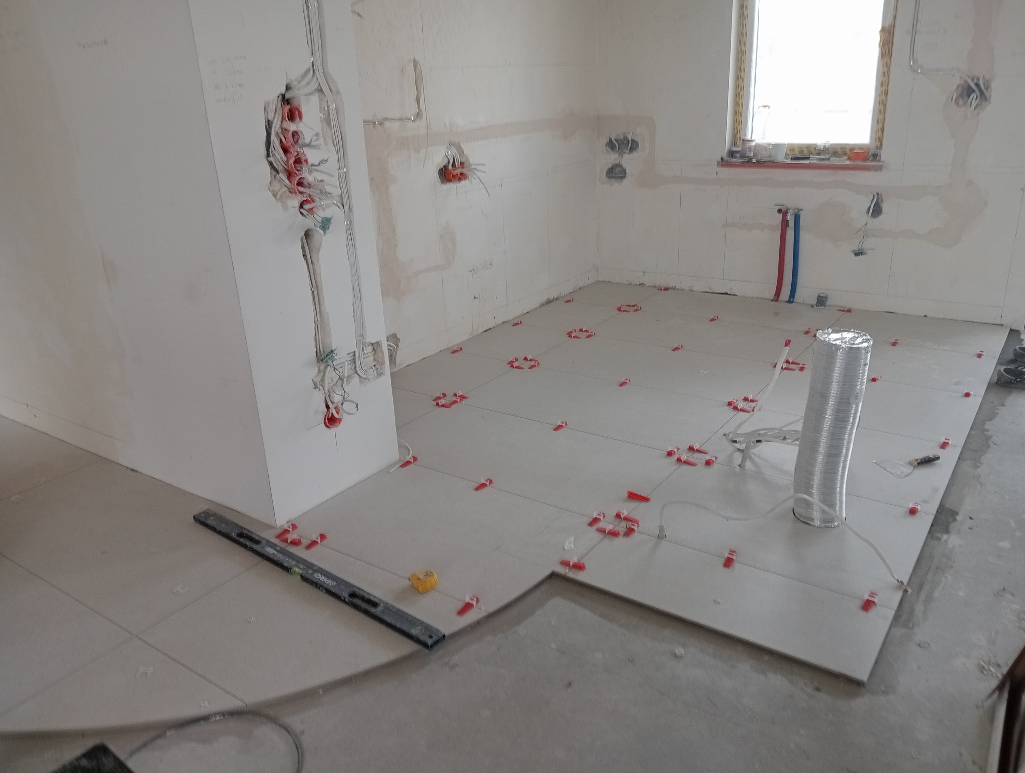 Jasne płytki podłogowe w trakcie układania, z czerwonymi klipsami poziomującymi. Widoczne przewody elektryczne i rury, przygotowane ściany. Poziomica i narzędzia glazurnicze.