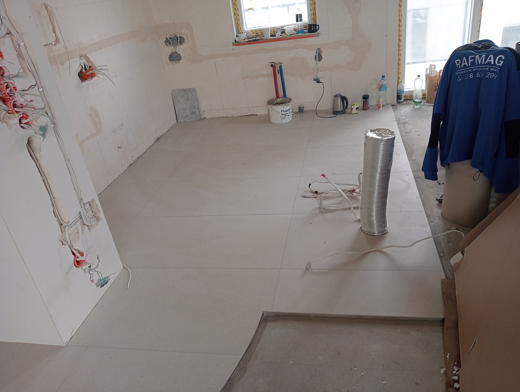 Jasne płytki podłogowe w trakcie układania, widoczne kable i puszka z klejem. Częściowo wykończona podłoga w pomieszczeniu z instalacją elektryczną na ścianach.